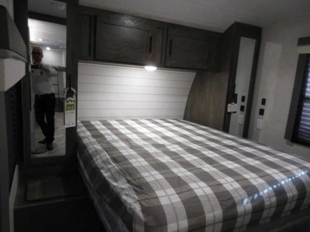 Interior of an RV rental bedroom with a comfortable plaid bed, overhead storage, and mirror reflection.. Forest River Wildwood 2021