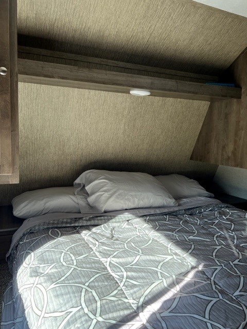 Sunlit RV rental bedroom features a bed with a gray patterned comforter, pillows, and an overhead shelf with a light.. Dutchmen Coleman Lantern 2018