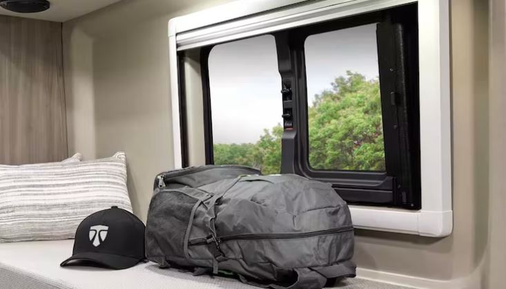 Experience comfort in an RV rental. Featuring a grey backpack, cap, and pillow by a window with a lush green view.. 2023 Thor Motor Coach Sprinter Tellaro 20j 20j 2023