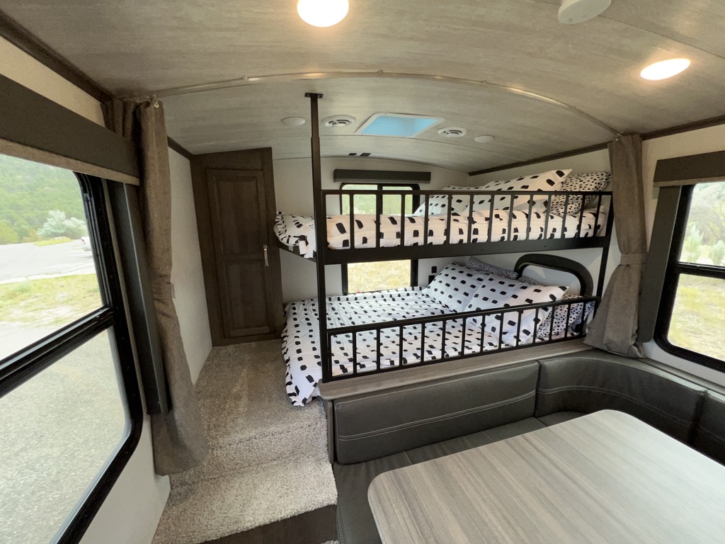 Interior of an RV rental showing modern bunk beds with spotted bedding, a dinette, and large windows offering scenic views.. Keystone RV Cougar 2022