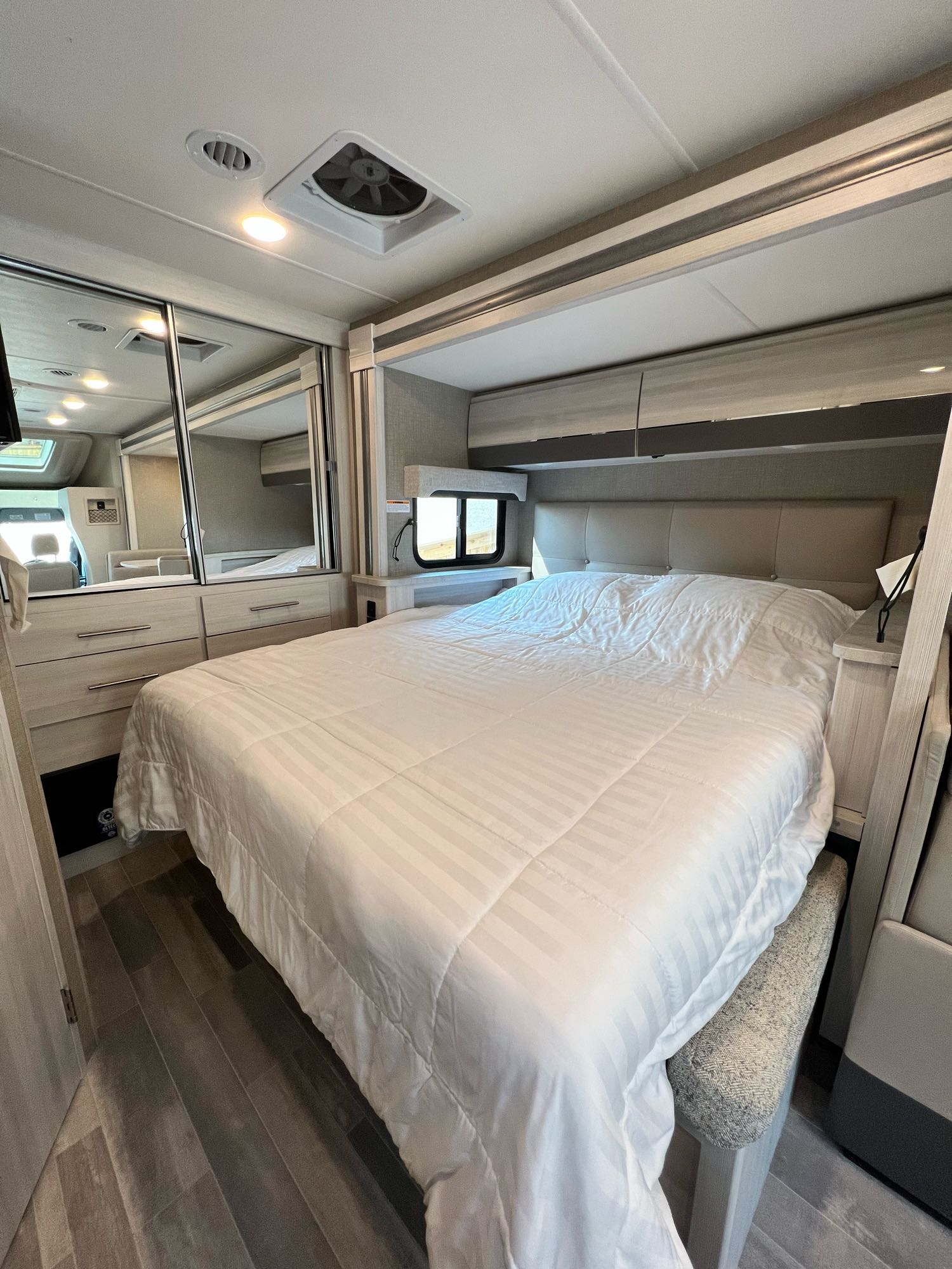 Spacious, modern RV rental bedroom features a comfortable bed with white linens, mirrored wardrobe, and overhead storage.. Thor Gemini 2023