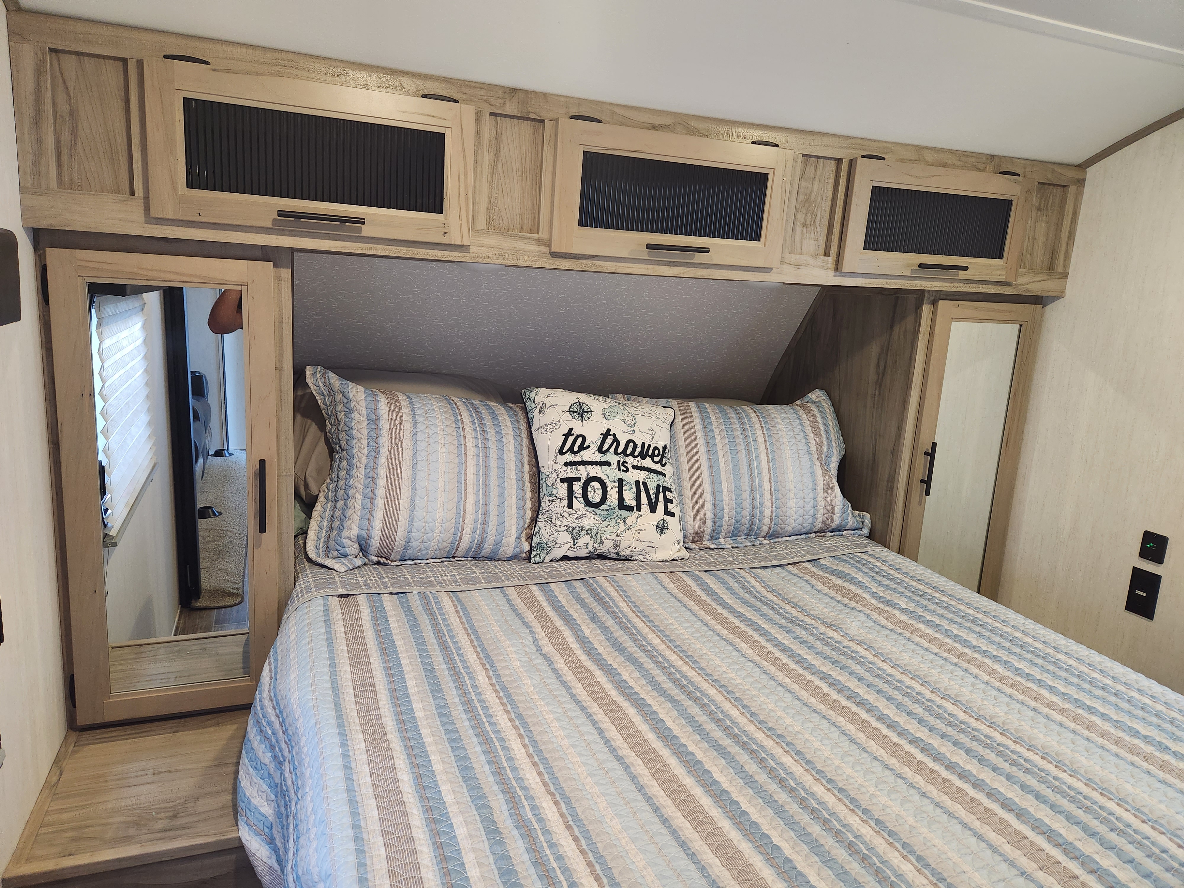 Cozy RV rental bedroom features a striped bed, 'to travel' pillow, overhead cabinets, and mirrored closets.. Forest River Cherokee Alpha Wolf 2022