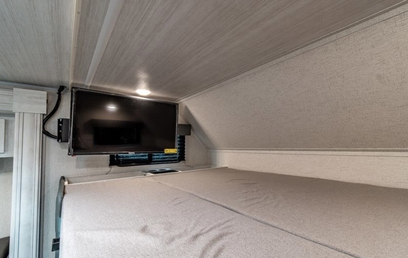 An RV rental's over-cab bunk bed with a wall-mounted TV, providing a compact and functional sleeping space.. Entrada East to West 2022