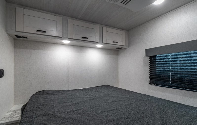 RV rental bedroom interior featuring overhead cabinets, bed with dark blanket, and window blinds. Clean, modern design.. Entrada East to West 2022