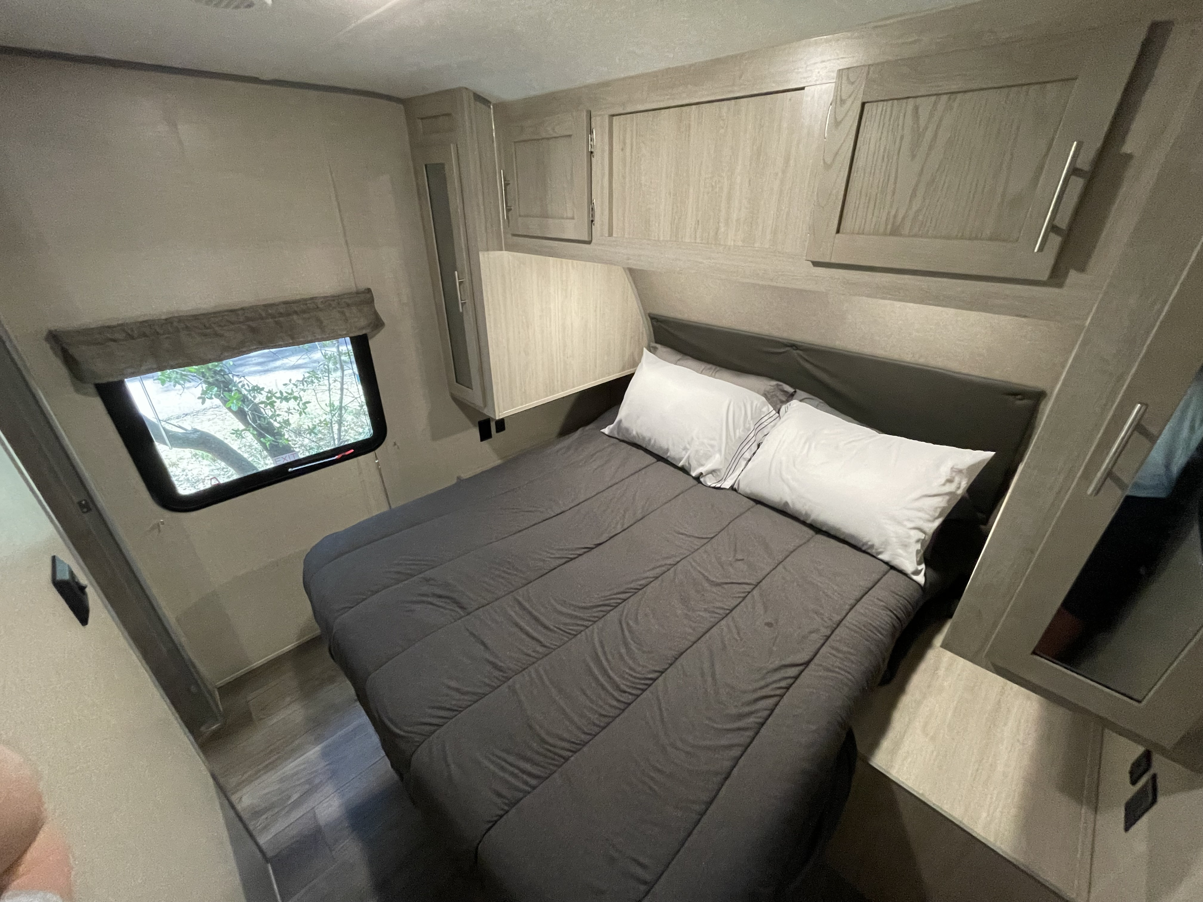 Cozy RV rental bedroom featuring a bed with dark grey bedding and white pillows, light wood cabinets, and a window with tree views.. Forest River Coachmen Catalina Legacy 2022