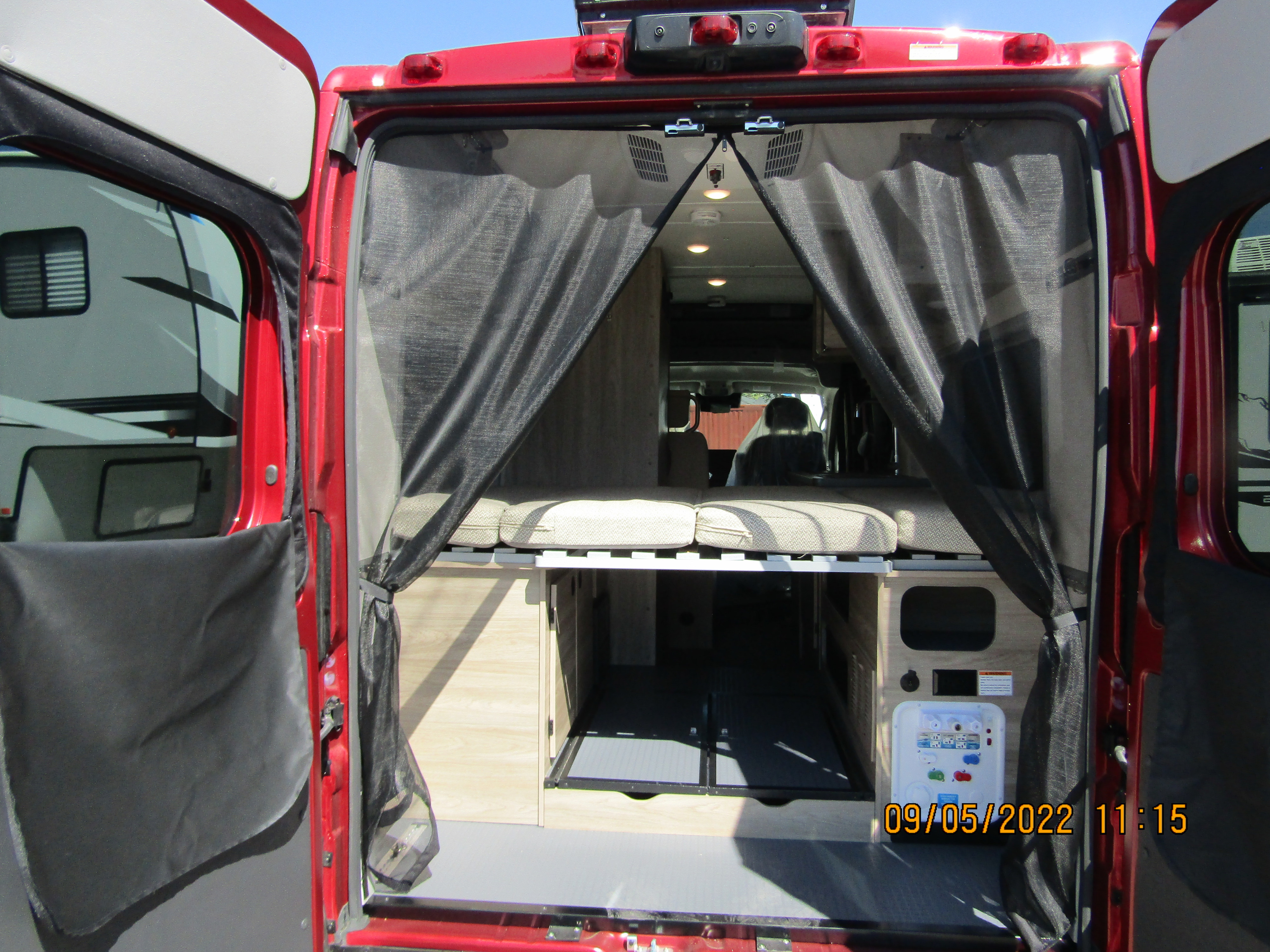 RV rental: Inside a red camper van with open rear doors, showing a bed, storage, utility panel, and mesh screens.. Winnebago SOLIS 2023