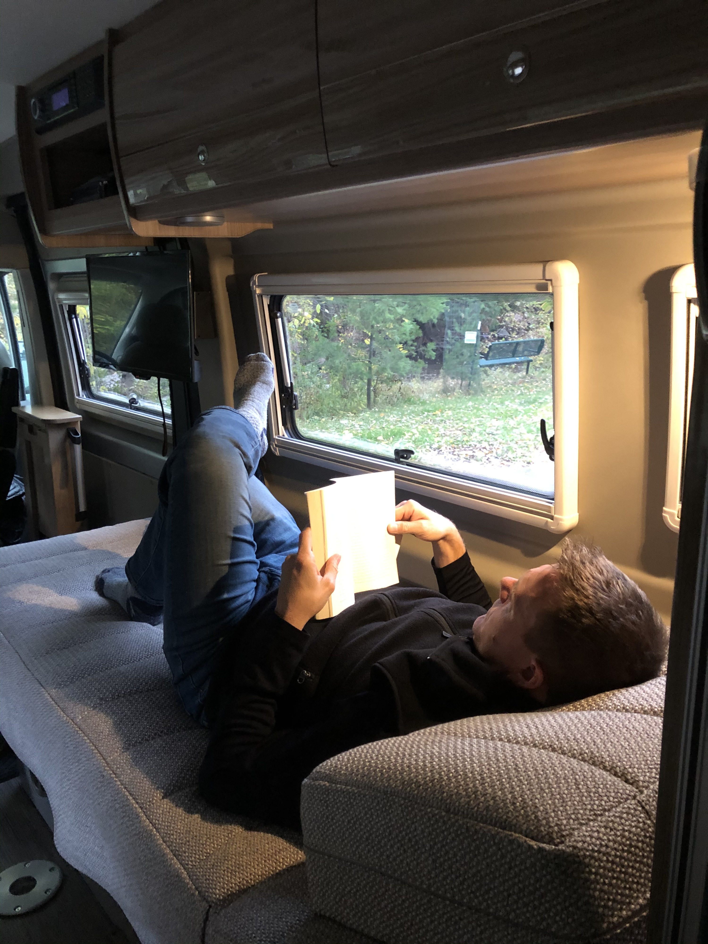 Relaxing in an RV rental: A person reads a book with feet up, enjoying the nature view from inside their cozy camper van.. Winnebago Travato 2019