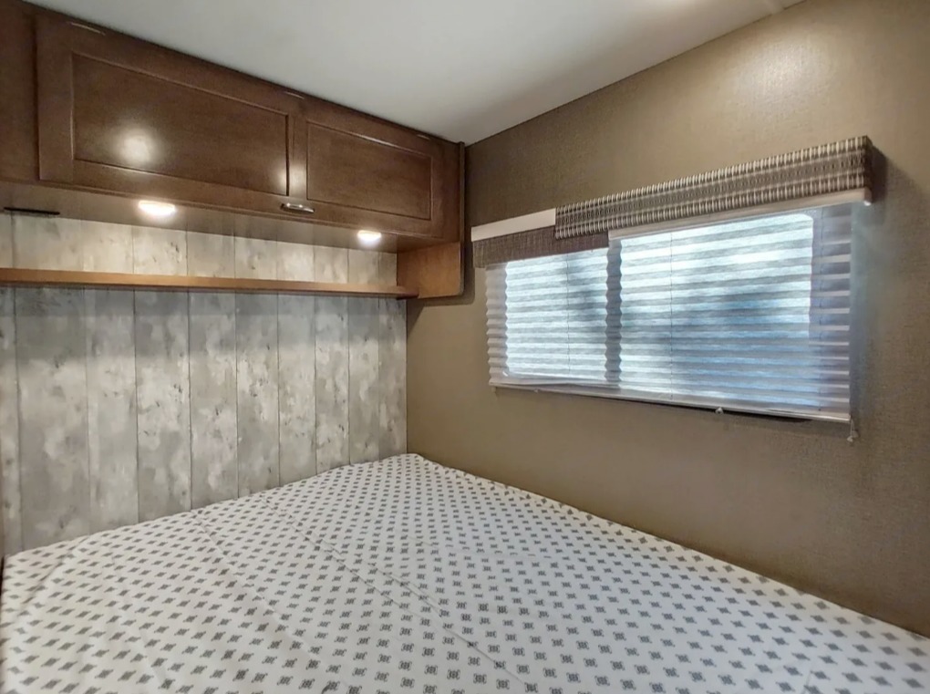 This RV rental bedroom offers a cozy bed, ample overhead cabinets, and a window with blinds for comfortable travels.. Winnebago Outlook 2019