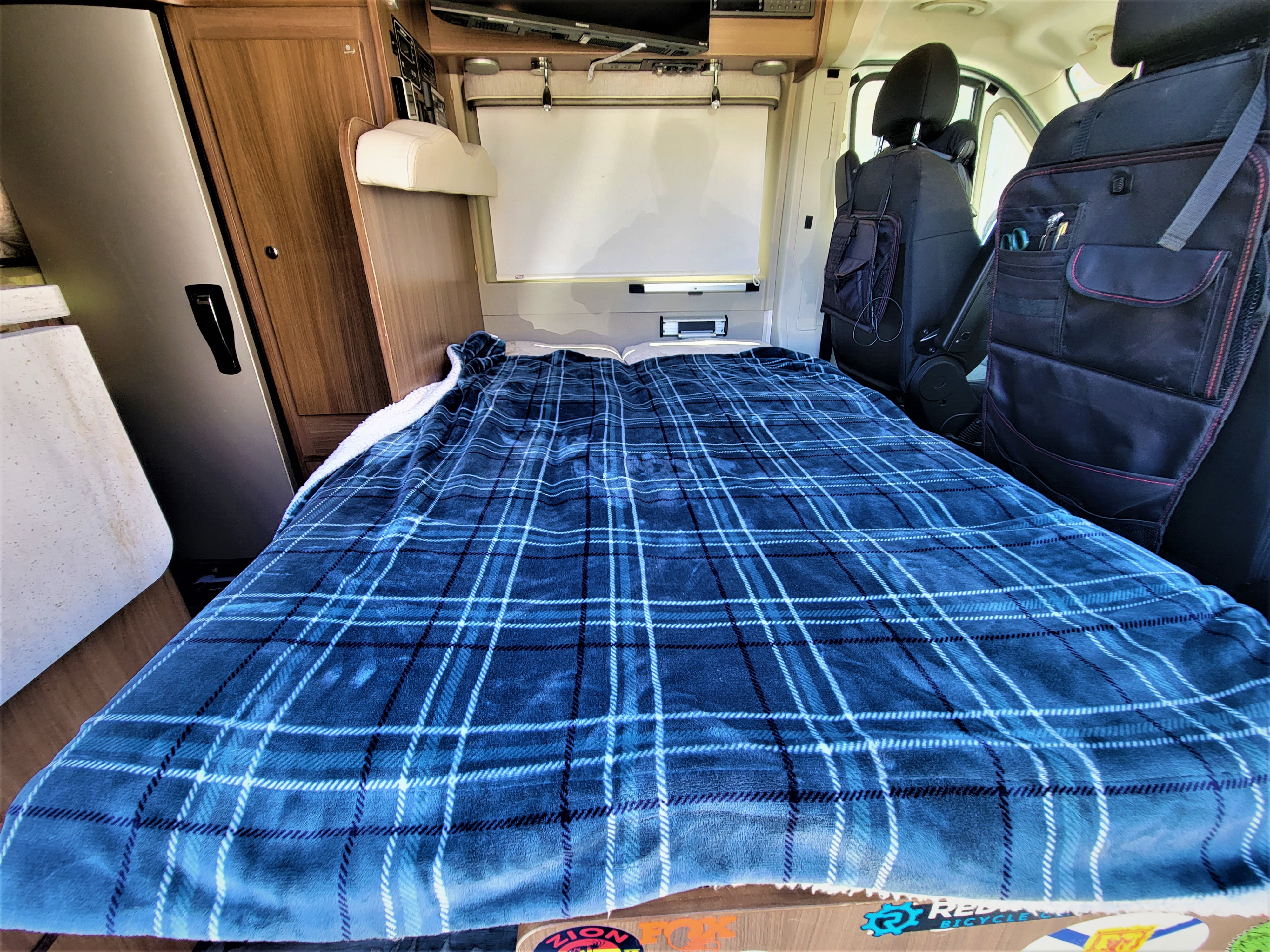 Cozy RV rental interior with a blue plaid bed, fridge, wooden cabinets, and front seat organizers, ready for travel.. Winnebago Travato 2018