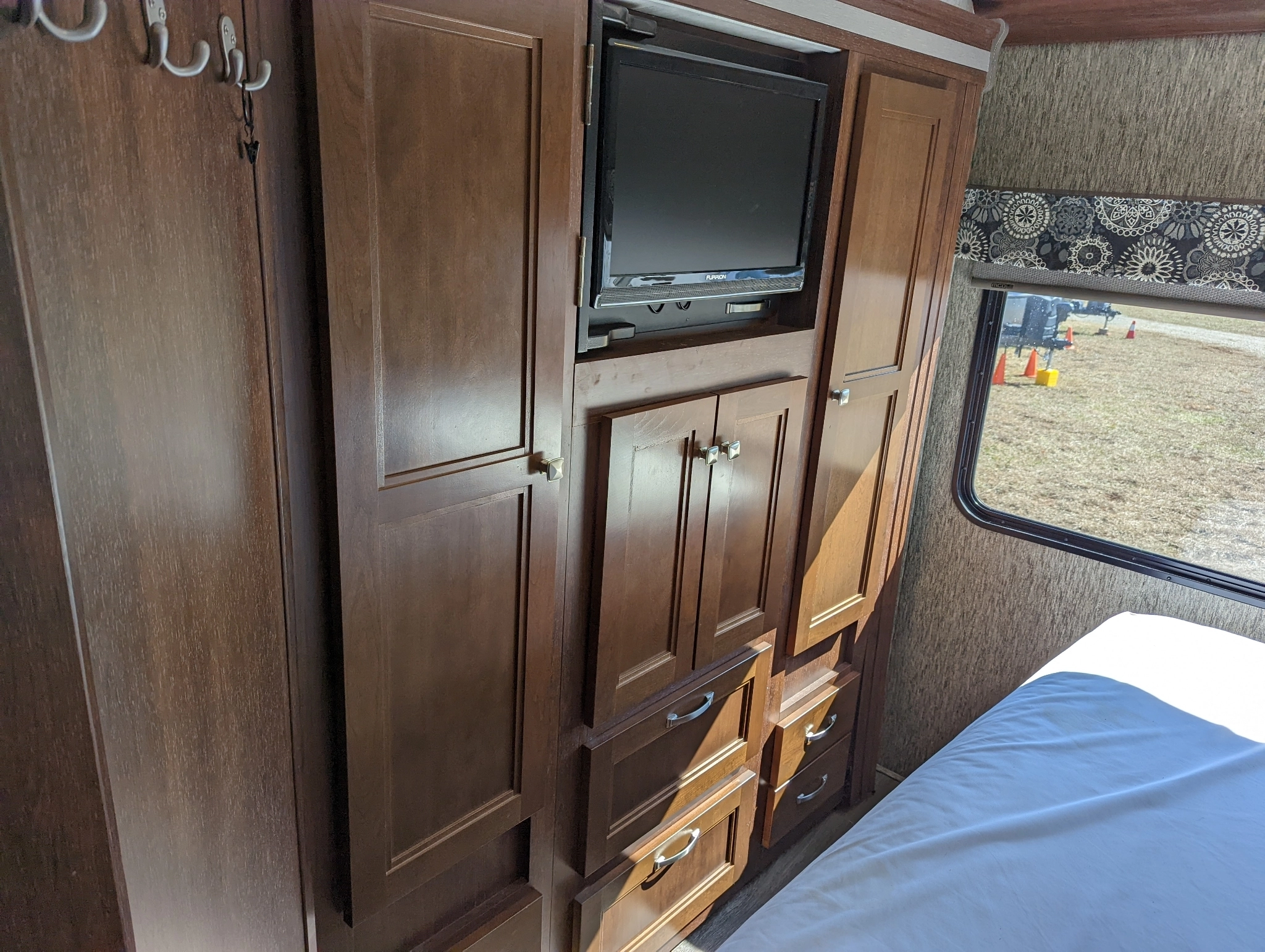 Explore this RV rental's organized bedroom, featuring dark wood storage, TV, and a window overlooking a grassy outdoor area.. Forest River Sunseeker 2018