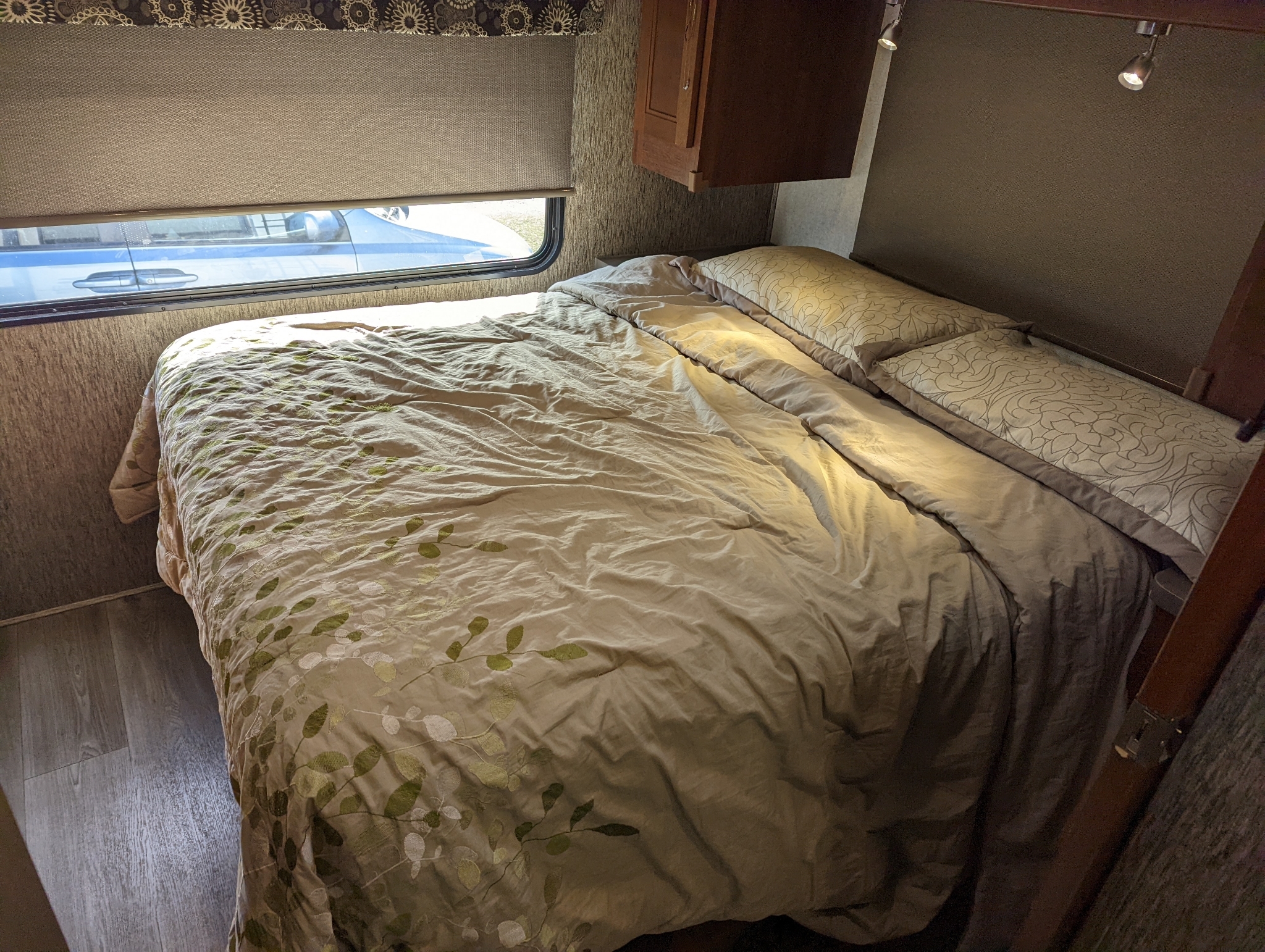 Inside a comfortable RV rental bedroom featuring a bed with leaf-patterned bedding, window, and overhead cabinet.. Forest River Sunseeker 2018