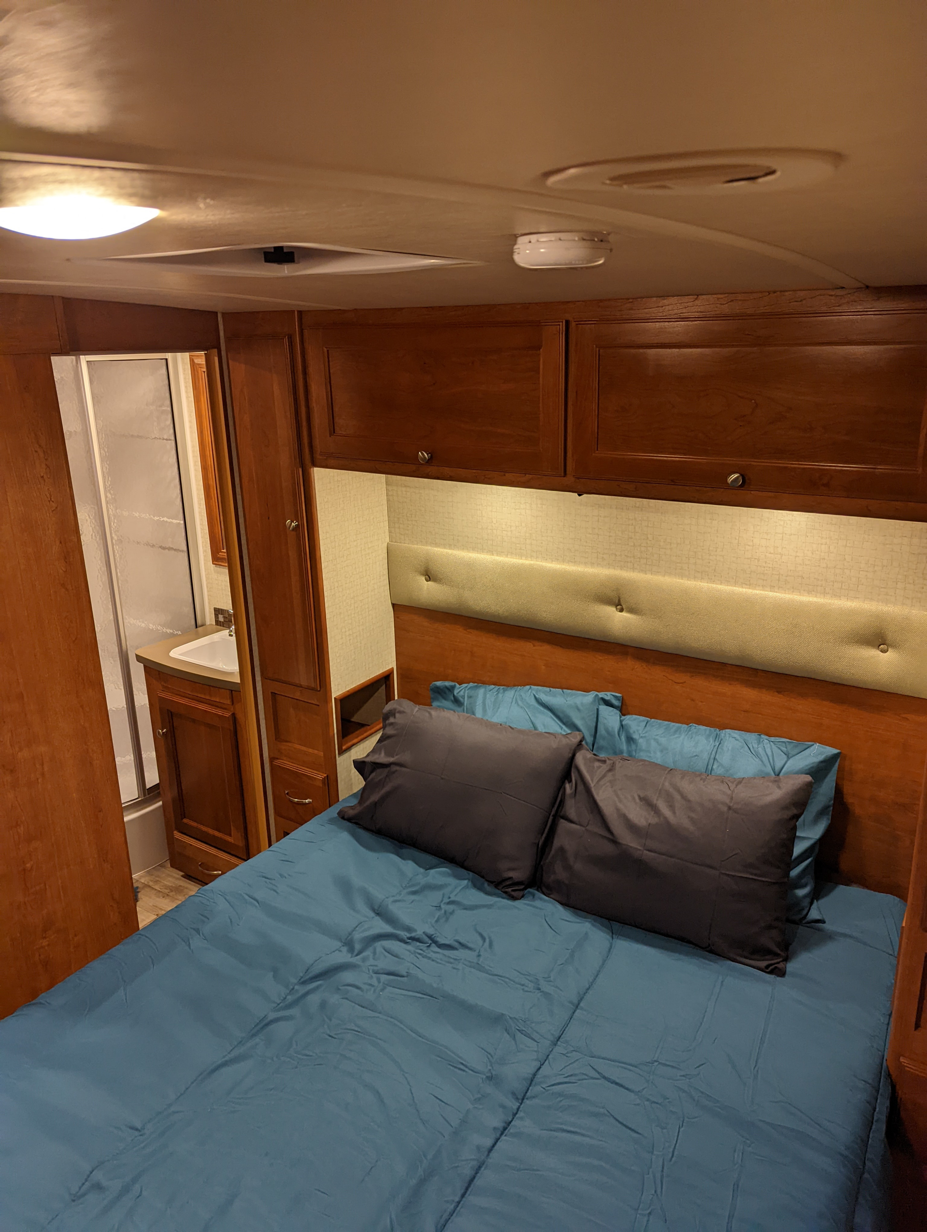 Cozy RV rental bedroom showing a bed with blue bedding, overhead wood cabinets, and an adjacent compact bathroom.. Winnebago Vista 2018