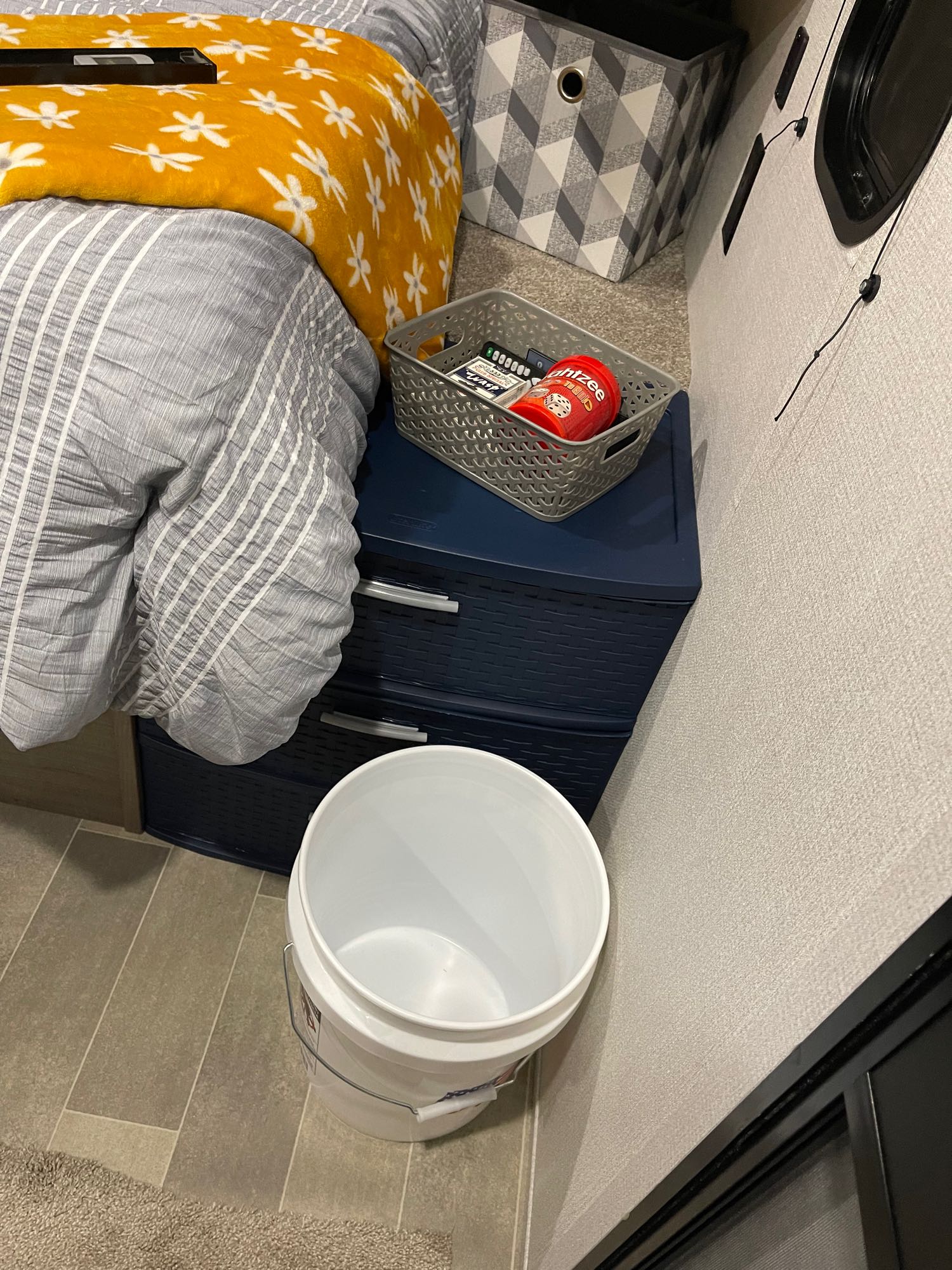 RV rental interior: A cozy bed with a yellow floral blanket, storage bins, games, and a bucket for practical use.. Keystone RV Hideout 2020