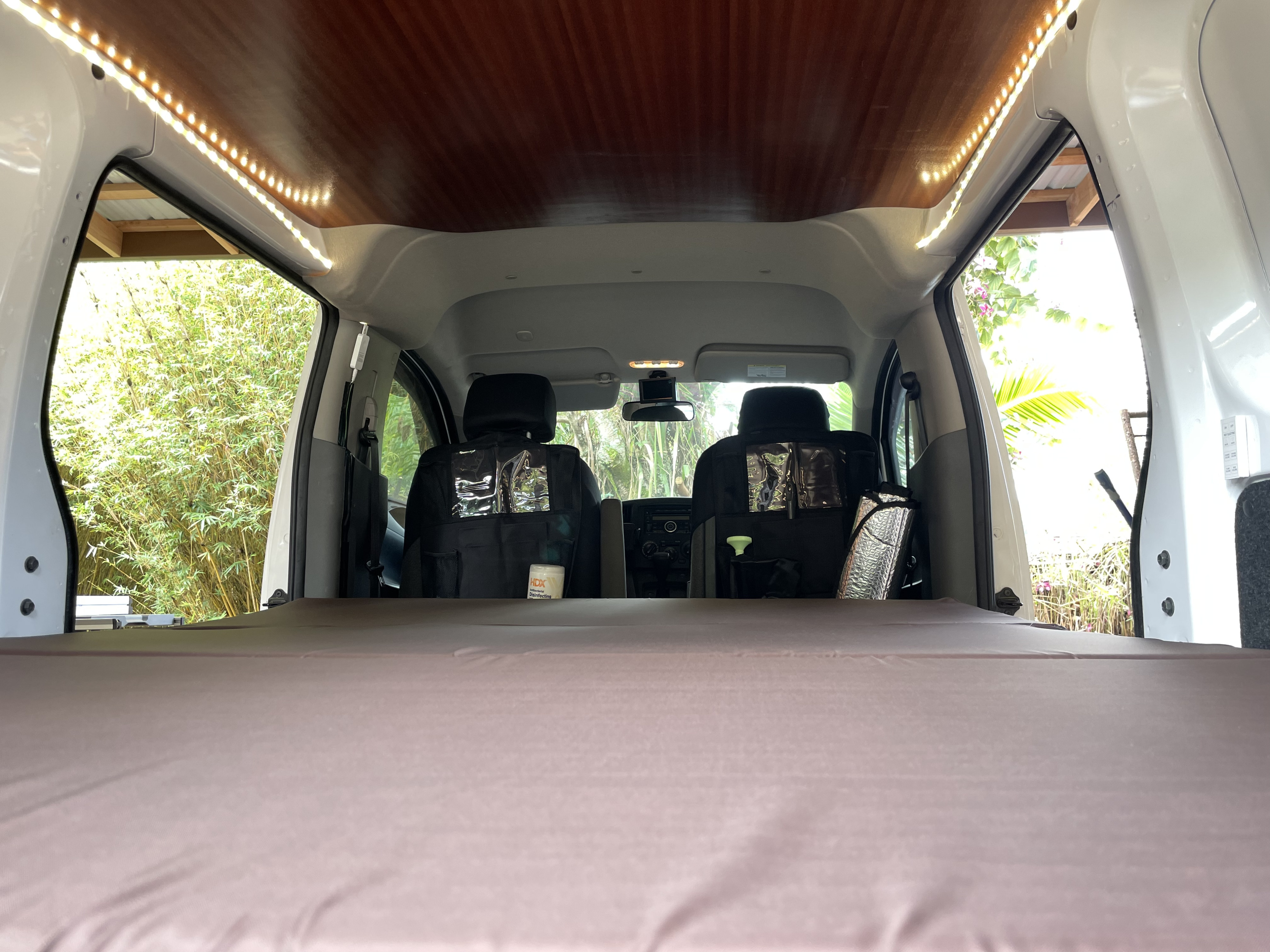 Inside an RV rental, a comfy bed and custom wooden ceiling with LED lights offer a cozy space looking out to nature.. Nissan NV200 2018