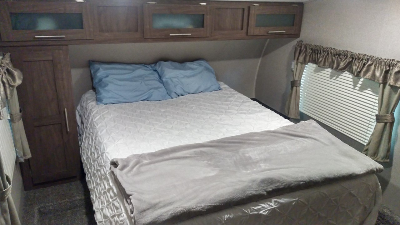 This comfortable RV rental bedroom features a neatly made bed, overhead storage, and a window with blinds.. Keystone Hideout 2019