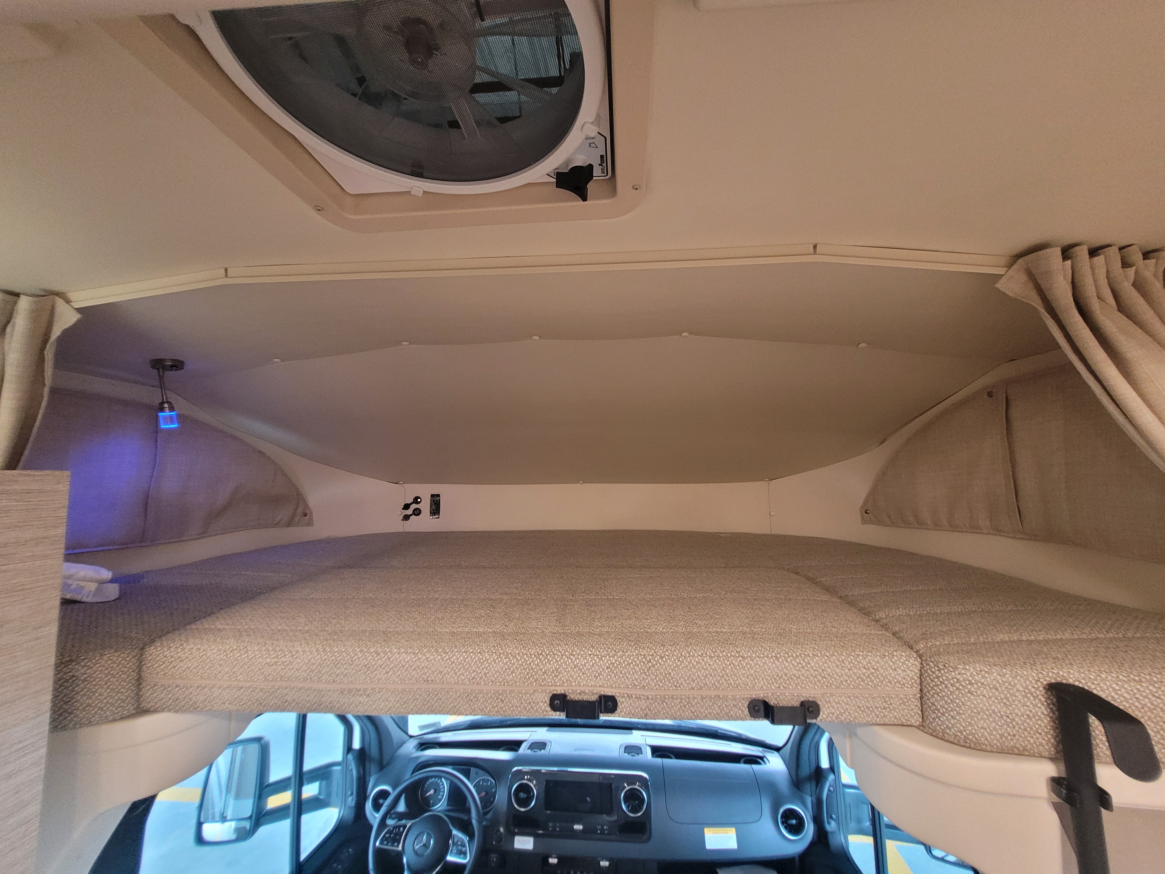 Cozy RV rental cab-over bunk with mattress, ceiling fan, and driver's cockpit visible below, ready for your road trip comfort.. Winnebago View 2023