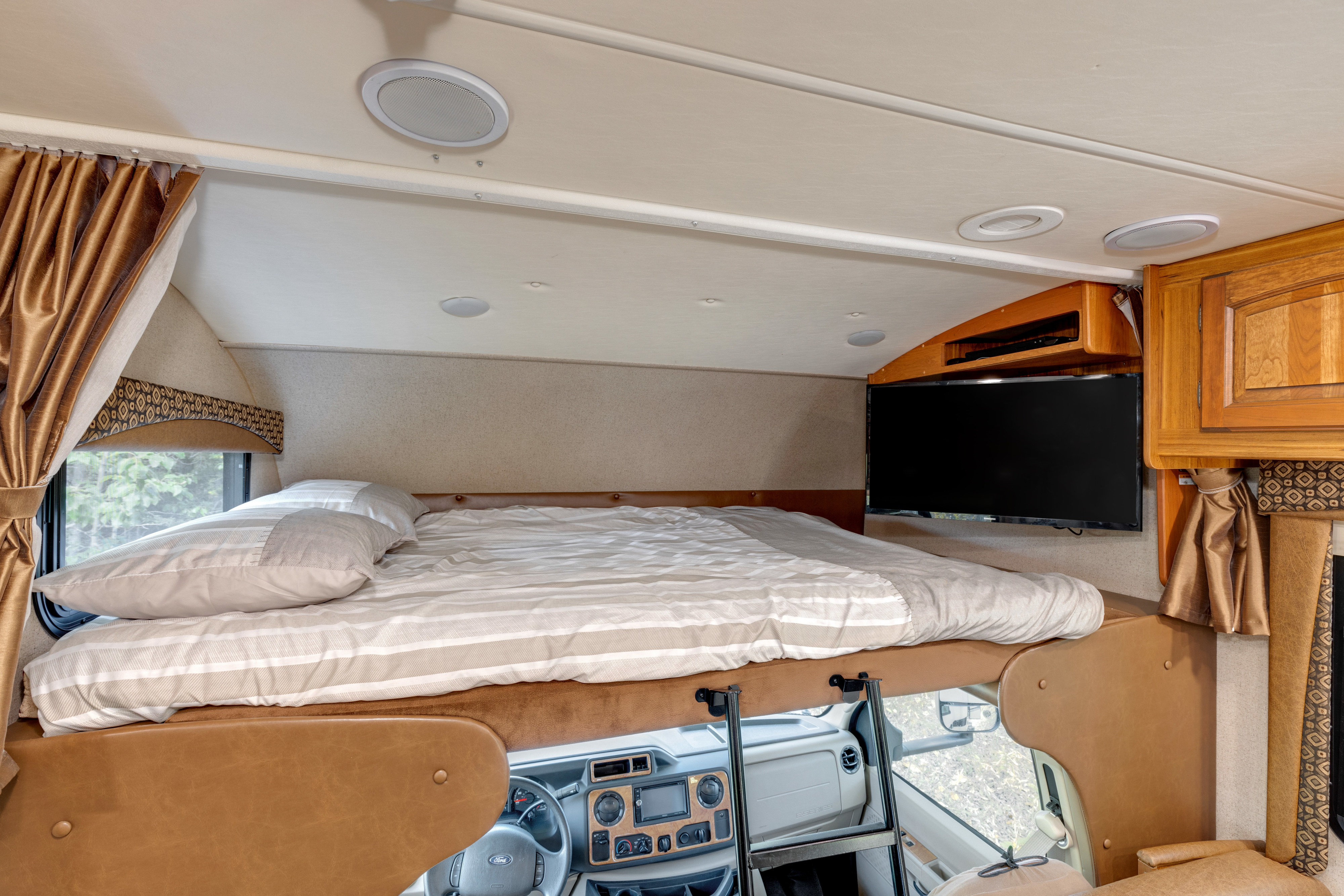Cozy RV rental cab-over bunk with striped bedding, a mounted TV, and dashboard view.. Jayco Greyhawk 2016