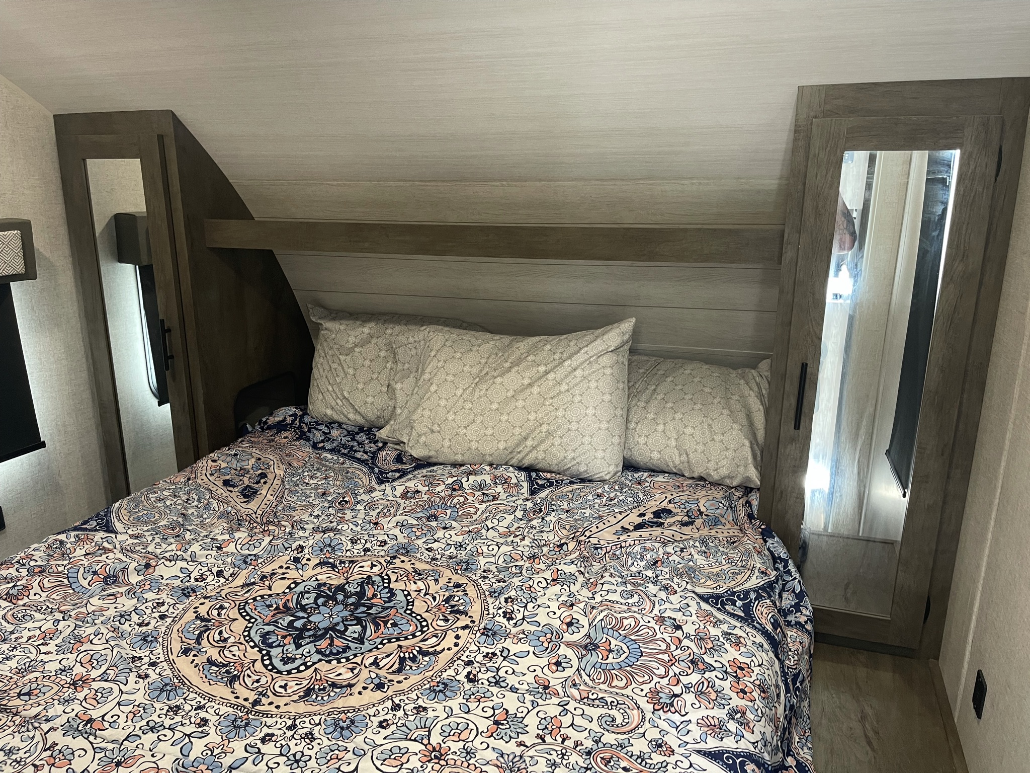 Cozy RV rental bedroom with a colorful patterned bed, mirrored wardrobes, and a wood-look headboard under a sloped ceiling.. Forest River Wildwood X-Lite 2020