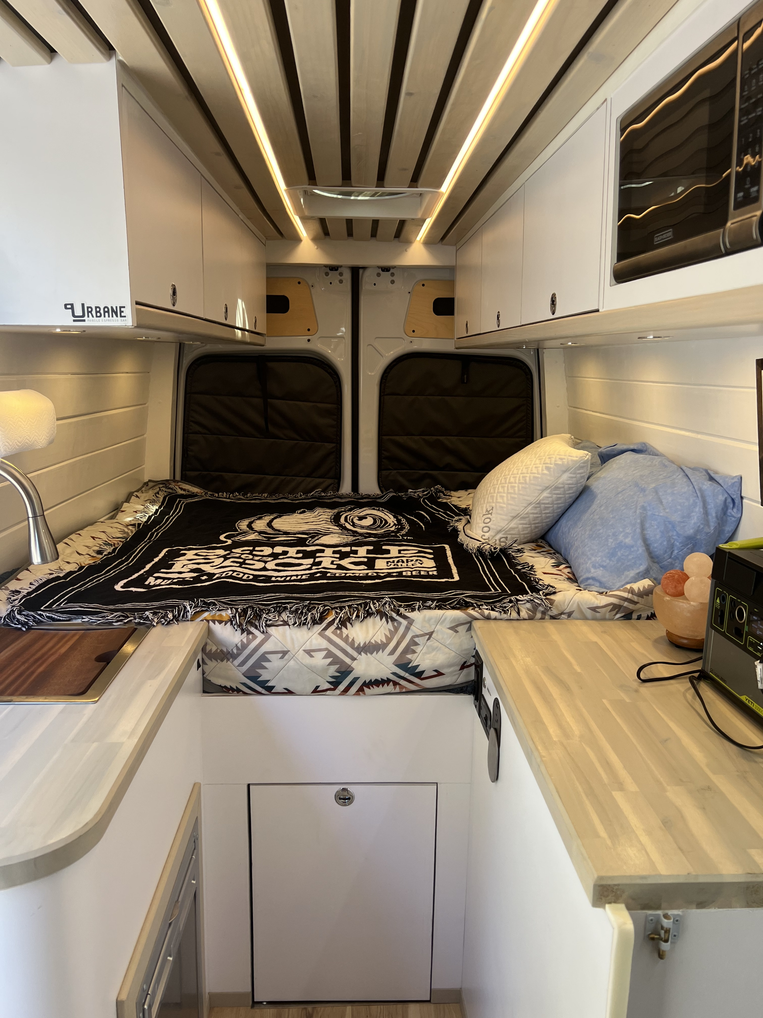 Bright rv rental interior: comfortable bed, kitchenette with sink, microwave, and overhead cabinets for convenient mobile living.. Mercedes-Benz Sprinter 2021