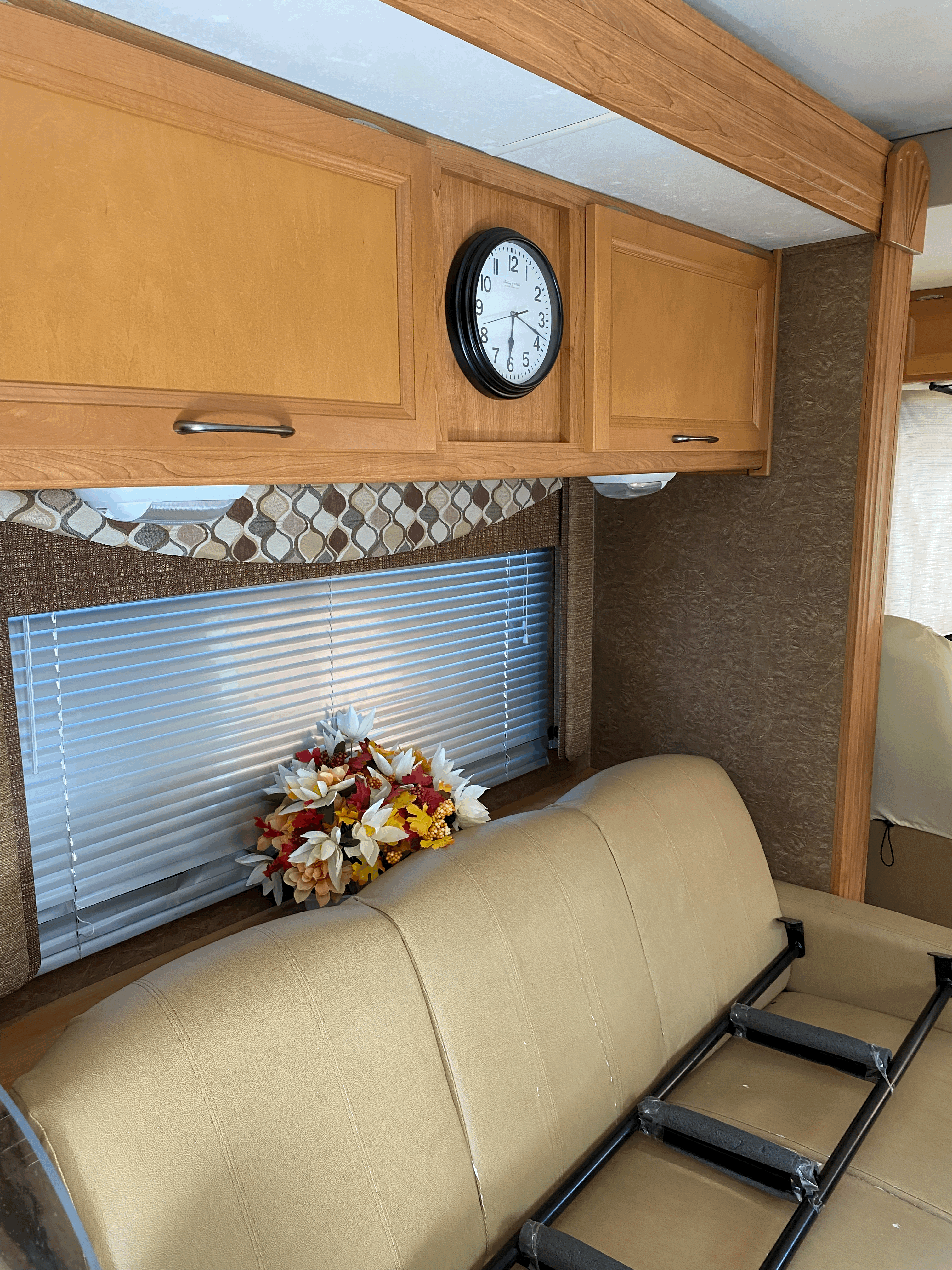 RV rental living area: light wood cabinets, a wall clock, window with blinds & flowers, and a tan sofa with a ladder.. Coachmen Pursuit 2015