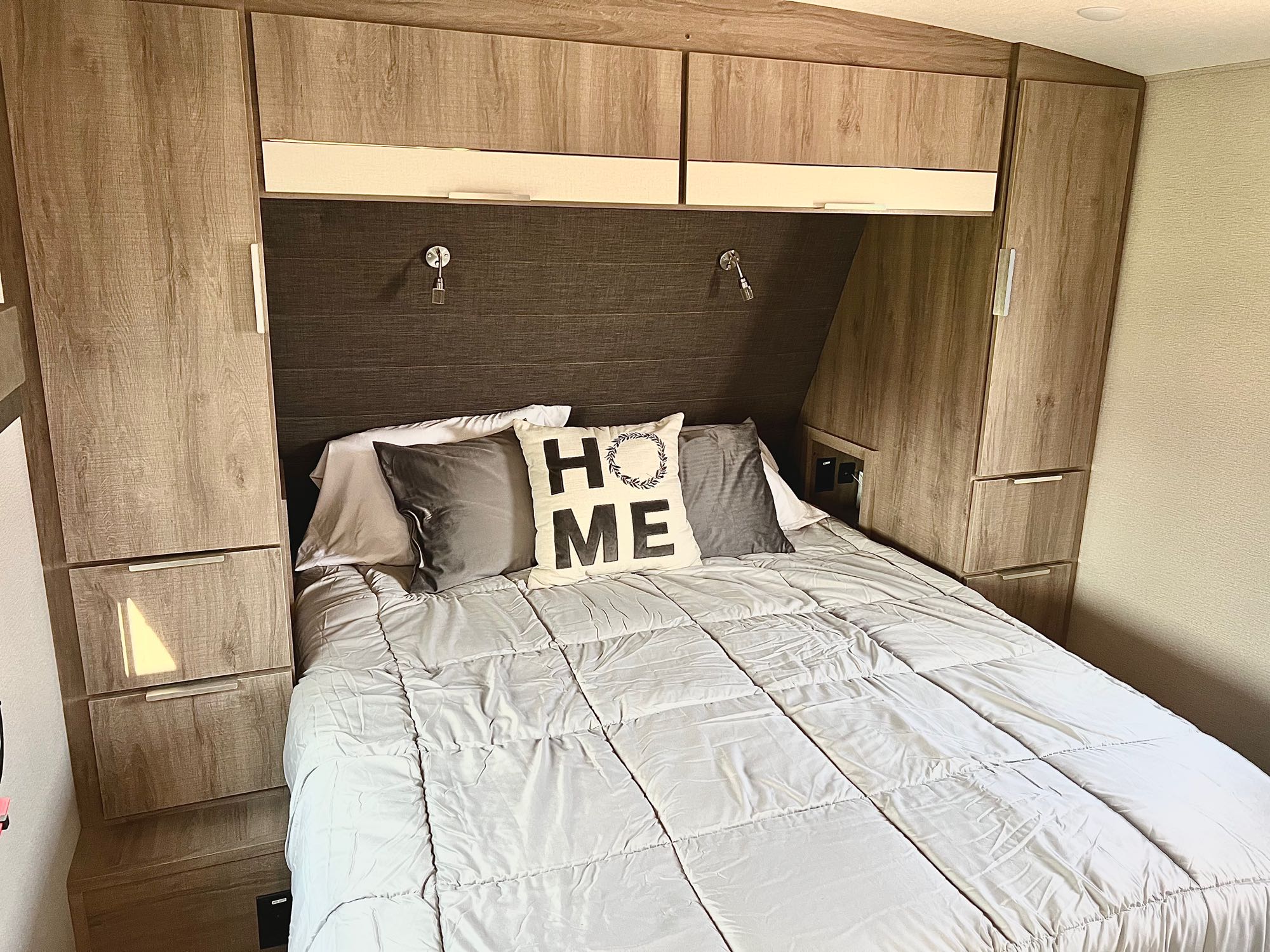 An RV rental bedroom setup, featuring a cozy bed with a "HOME" pillow, overhead cabinets, and side storage.. Grand Design Imagine 2021