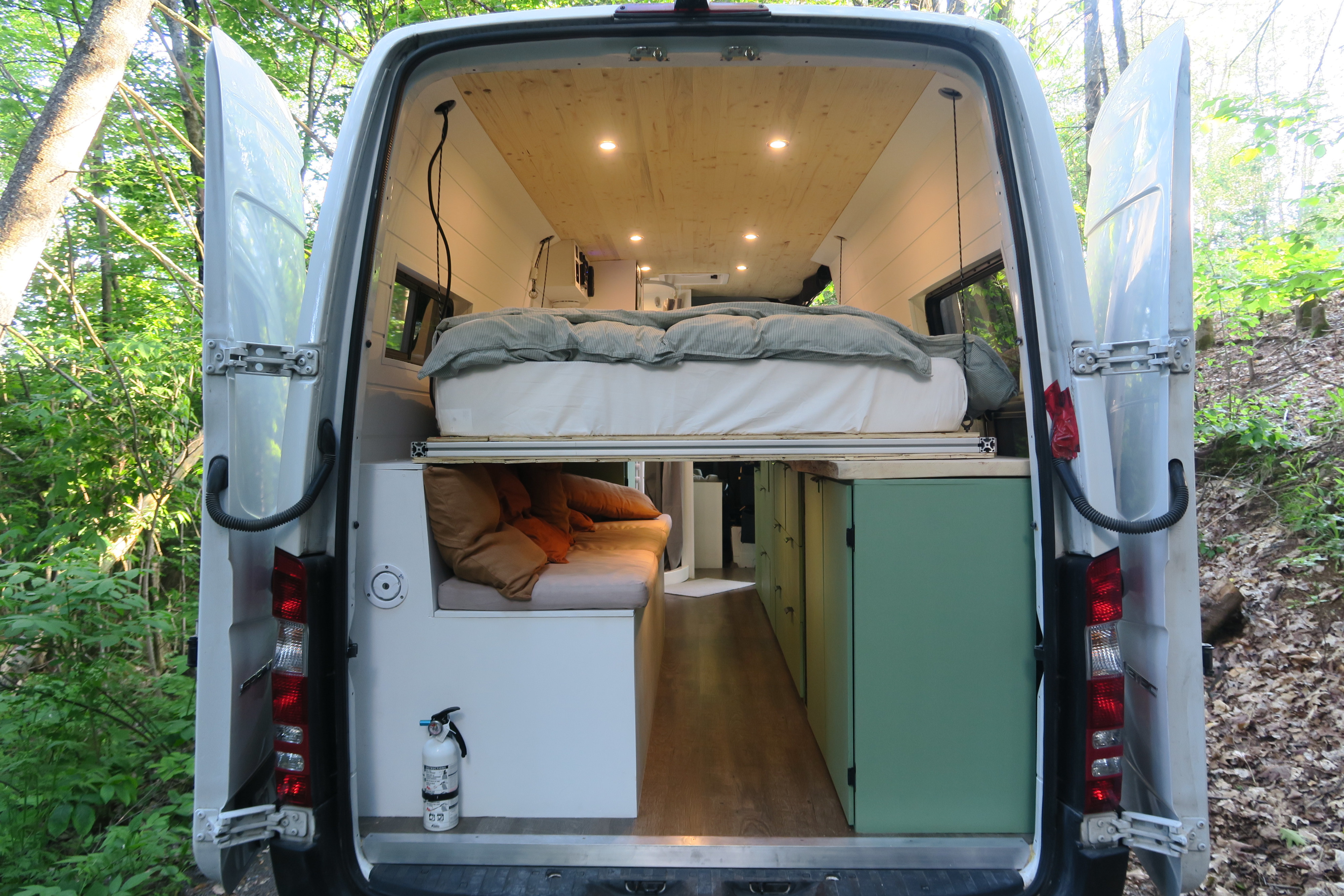 This cozy RV rental showcases a camper van's interior with a raised bed, bench lounge, and green cabinets, set in nature.. Other Sprinter 2016