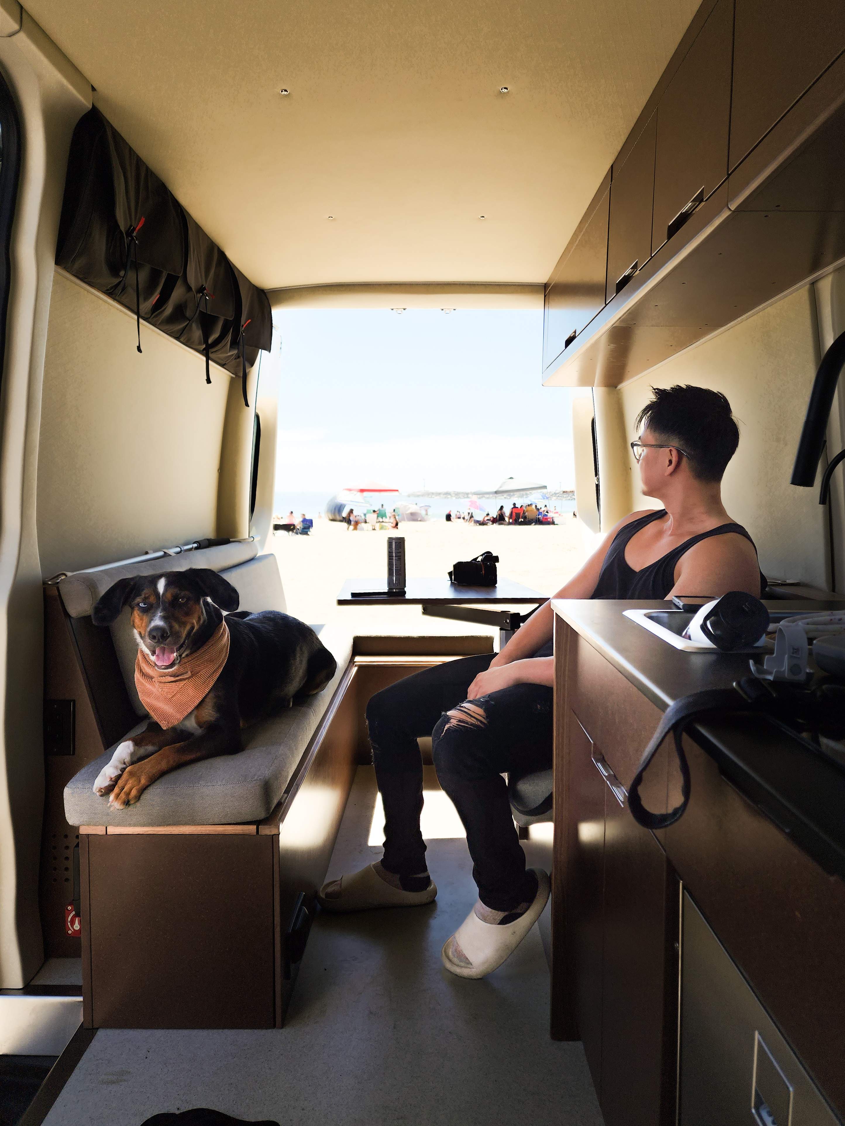 Inside an RV rental: A man and dog enjoy beach views. Overhead storage & kitchen are visible, with people on the sand outside.. Mercedes Sprinter 2021