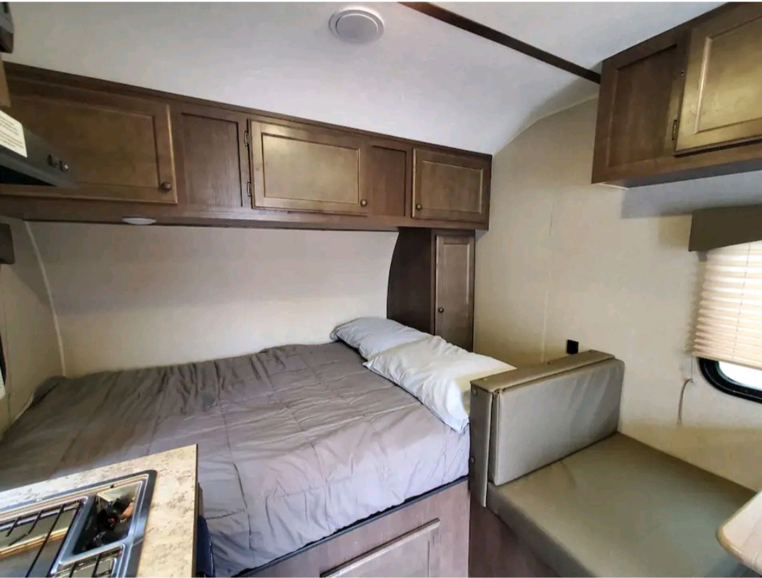 Cozy RV rental bedroom setup with a bed, overhead wood cabinets, a compact kitchenette, and a sofa/dinette area.. Sunset Park RV Sun Lite 2022