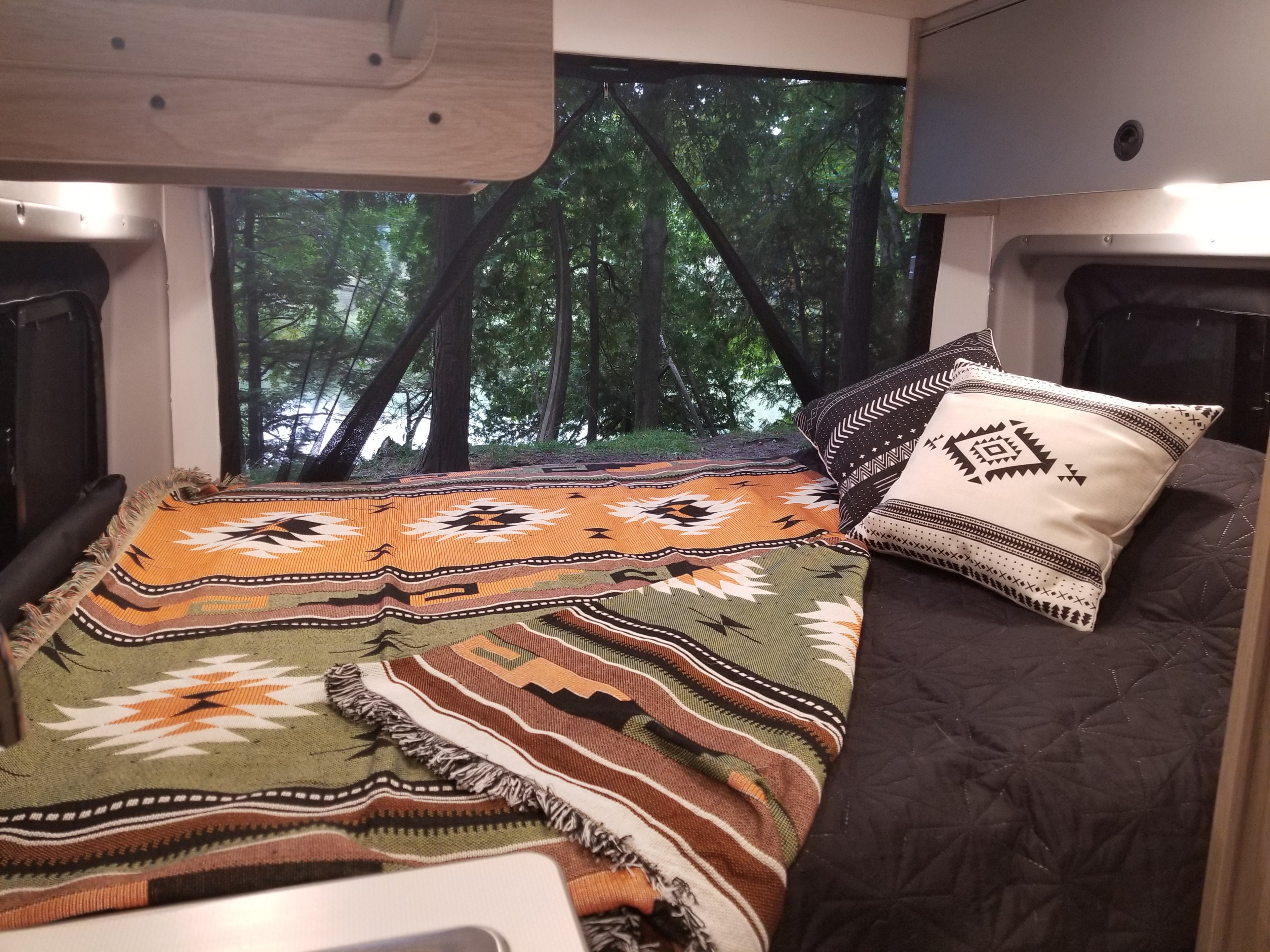 An inviting RV rental interior featuring a cozy bed with decorative blankets and pillows, offering a scenic forest and lake view.. Winnebago Solis 2022