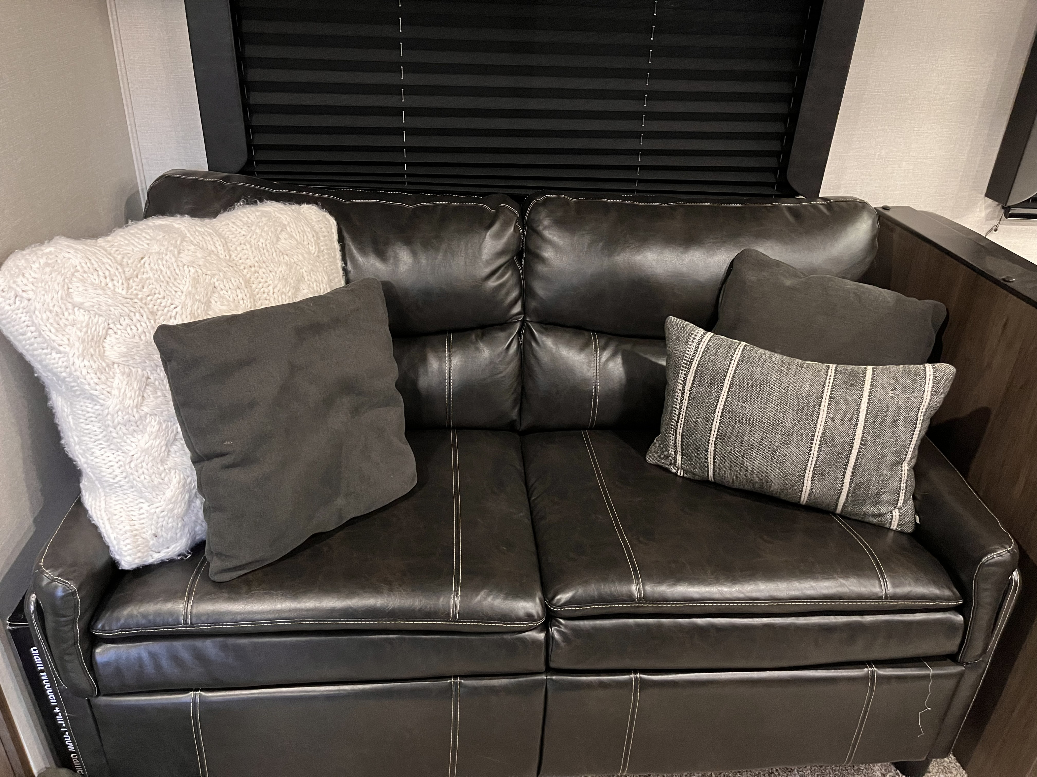 Dark brown sofa with white stitching, decorative knitted and striped pillows, and a black blind in an RV rental.. Forest River Tracer 2021