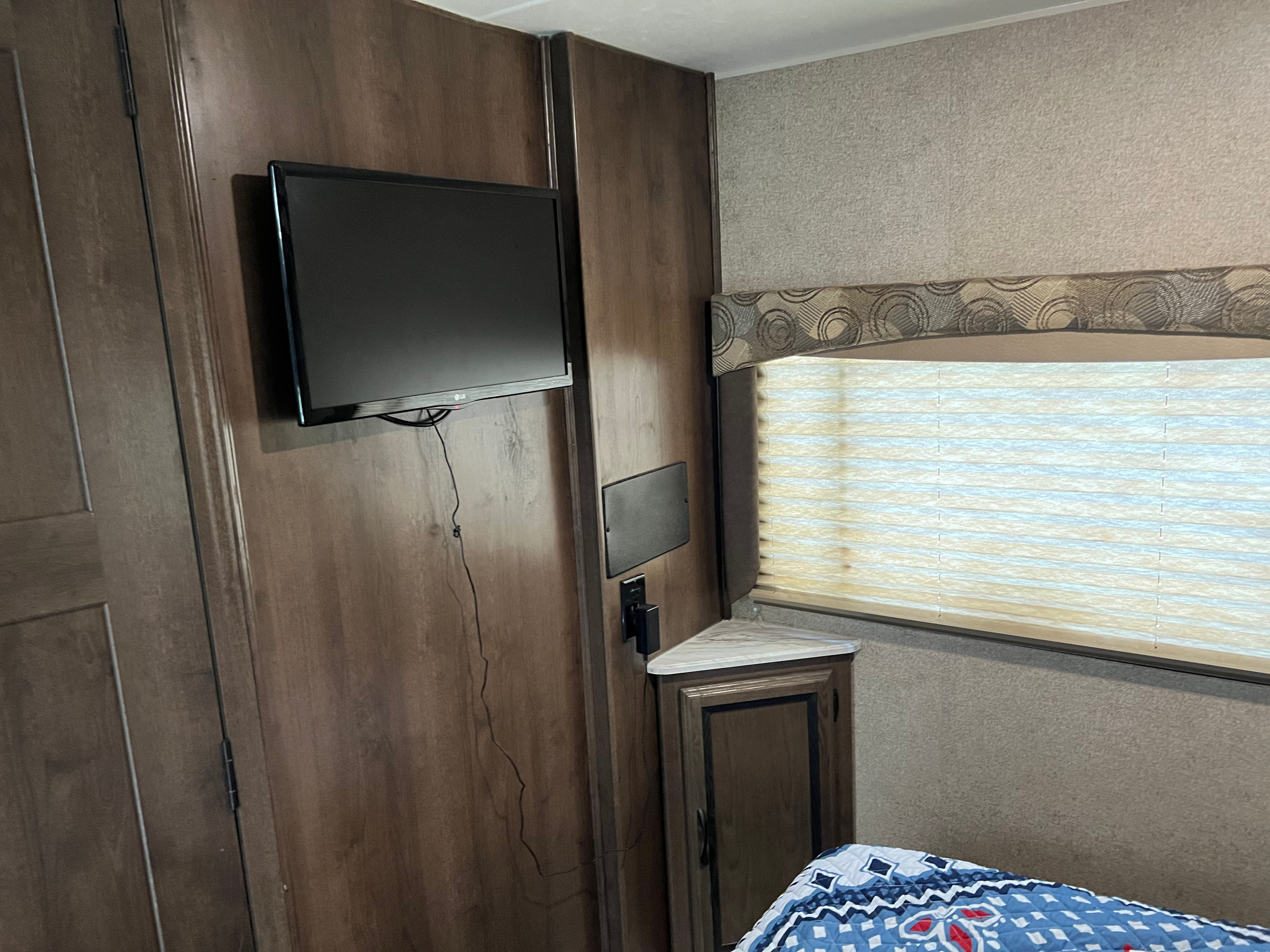RV rental bedroom with wall-mounted TV, window & blinds, and corner cabinet. Wood walls and patterned textiles visible.. Forest River Coachmen Freelander 2019