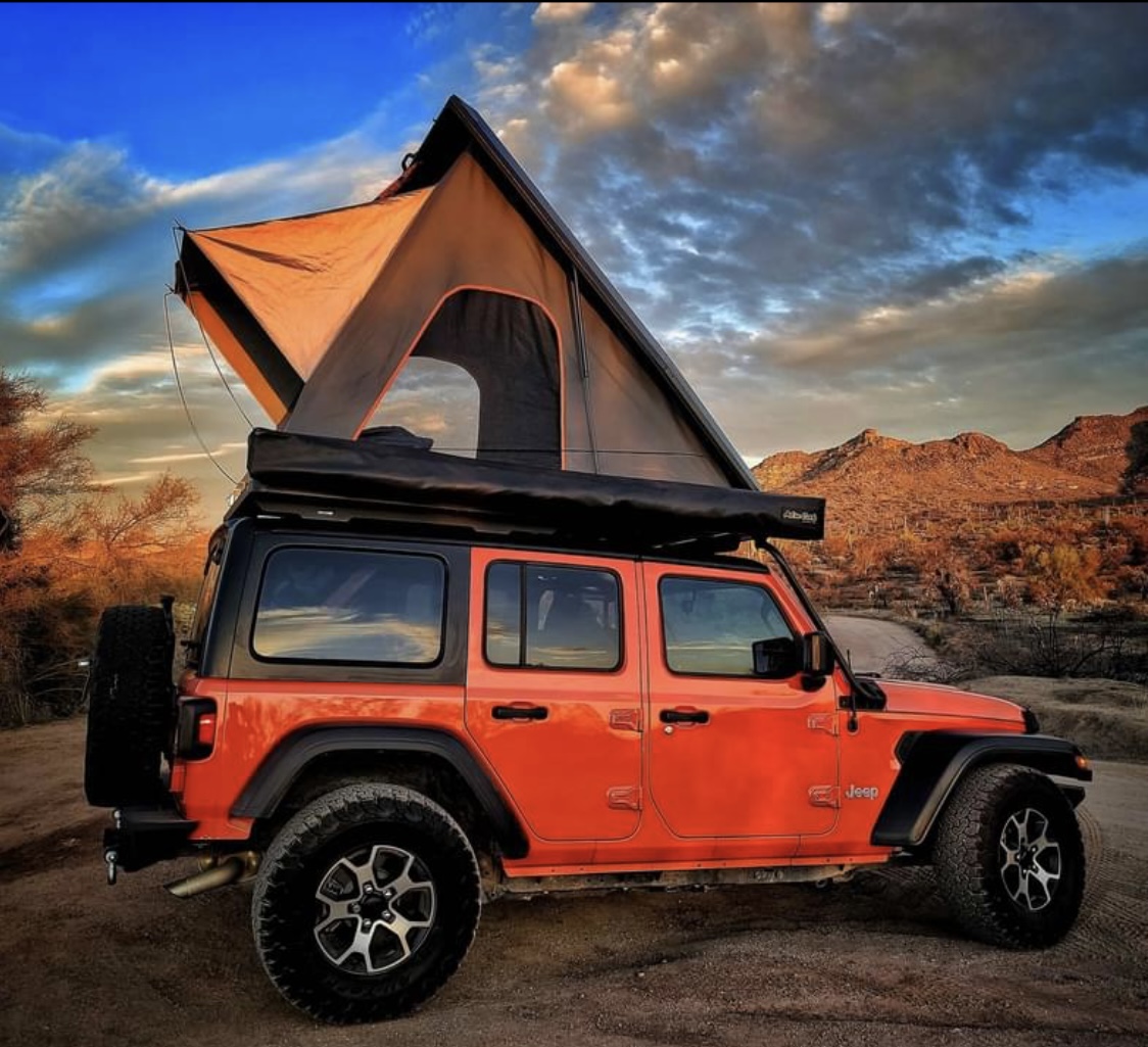 This vibrant orange Jeep Wrangler, equipped with a rooftop tent, offers an exciting RV rental experience in a rugged desert landscape.. Jeep Wrangler 2018