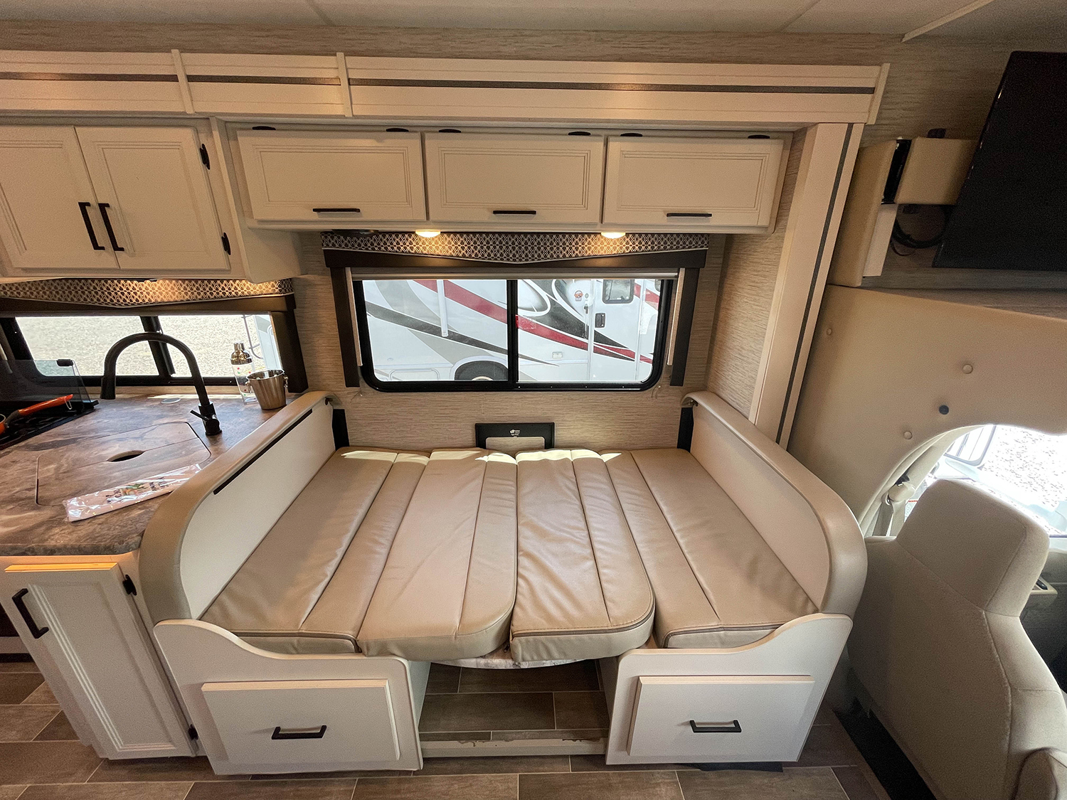 Inside an RV rental: a flexible dinette converts to a bed, beside a compact kitchen area. Overhead cabinets & front cab visible.. Thor Chateau 2022