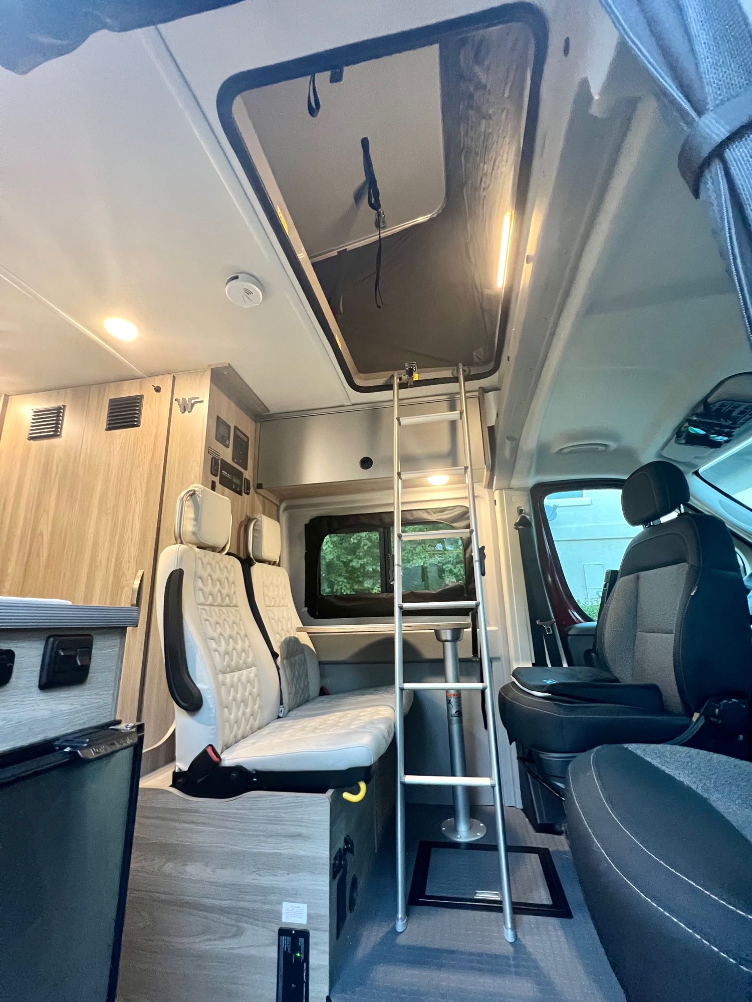 Inside an RV rental: a ladder to the pop-top bed, cream seating, wood cabinetry with fridge, and front cockpit view.. Winnebago Solis 2022