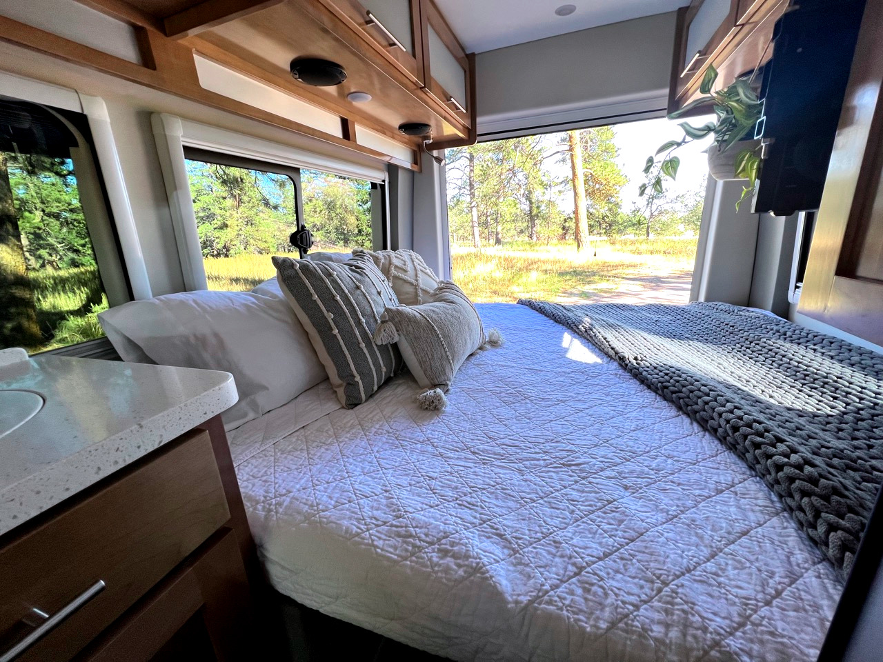 Cozy RV rental interior: comfortable bed with white bedding, decorative pillows & knit blanket, offering serene forest views outside.. Coachmen Beyond 22D 2020