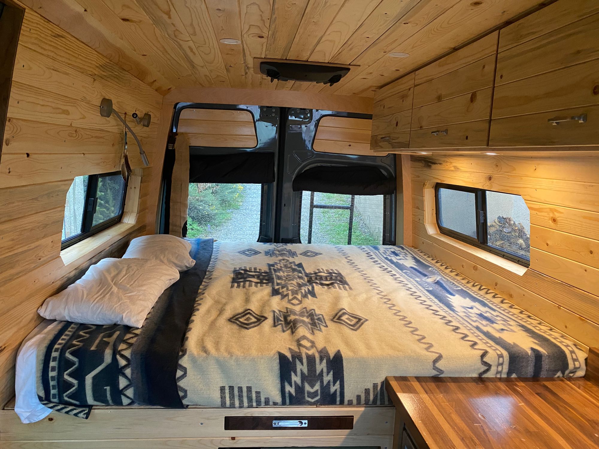 Cozy wood-paneled RV rental camper van interior: Bed with patterned blanket, built-in storage, and rear view.. Mercedes-Benz Sprinter 2021
