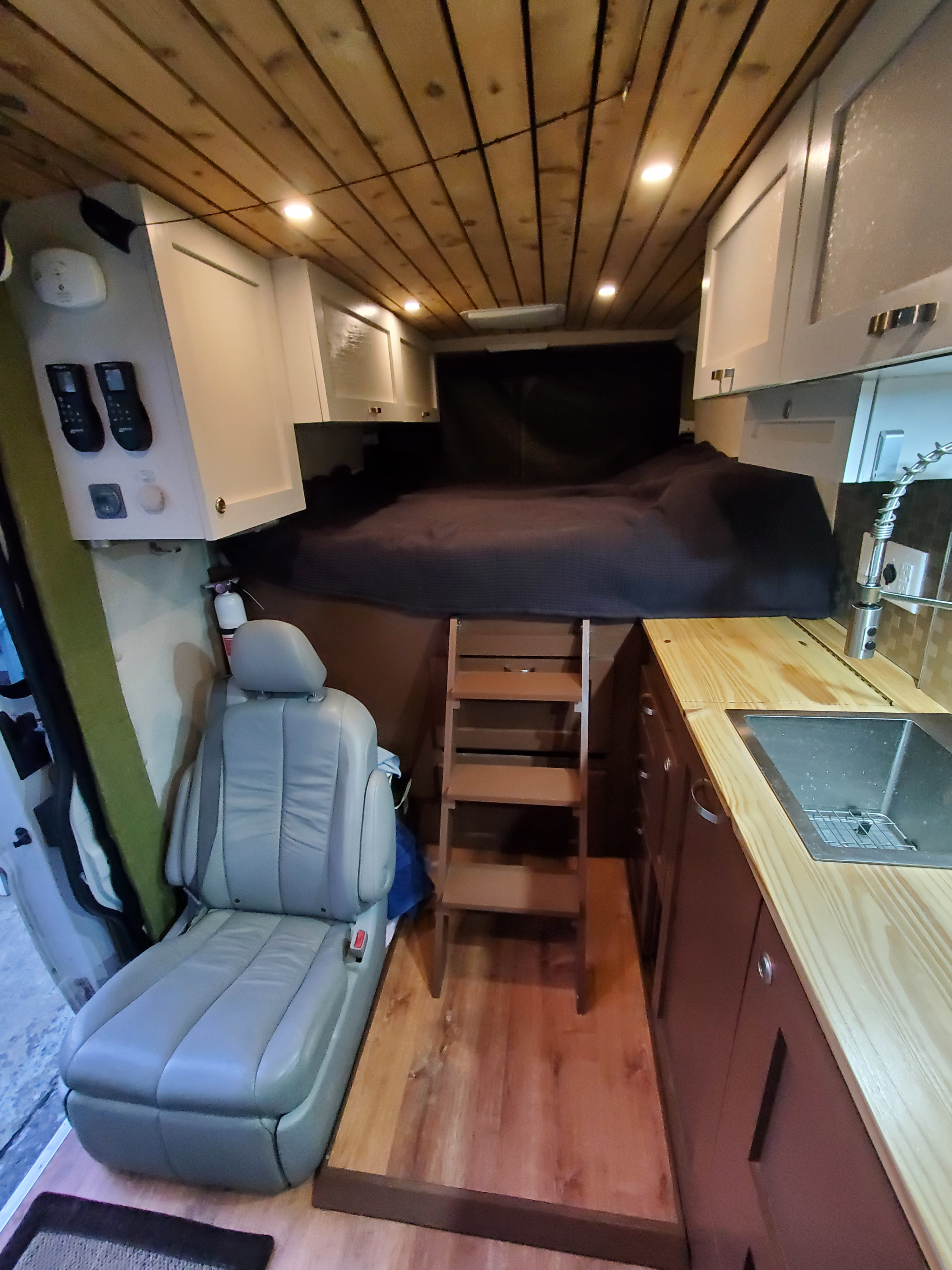 Inside a modern rv rental van: a cozy raised bed with ladder, compact kitchen with sink, light wood ceiling, and comfortable seating.. Custom Camper 2017
