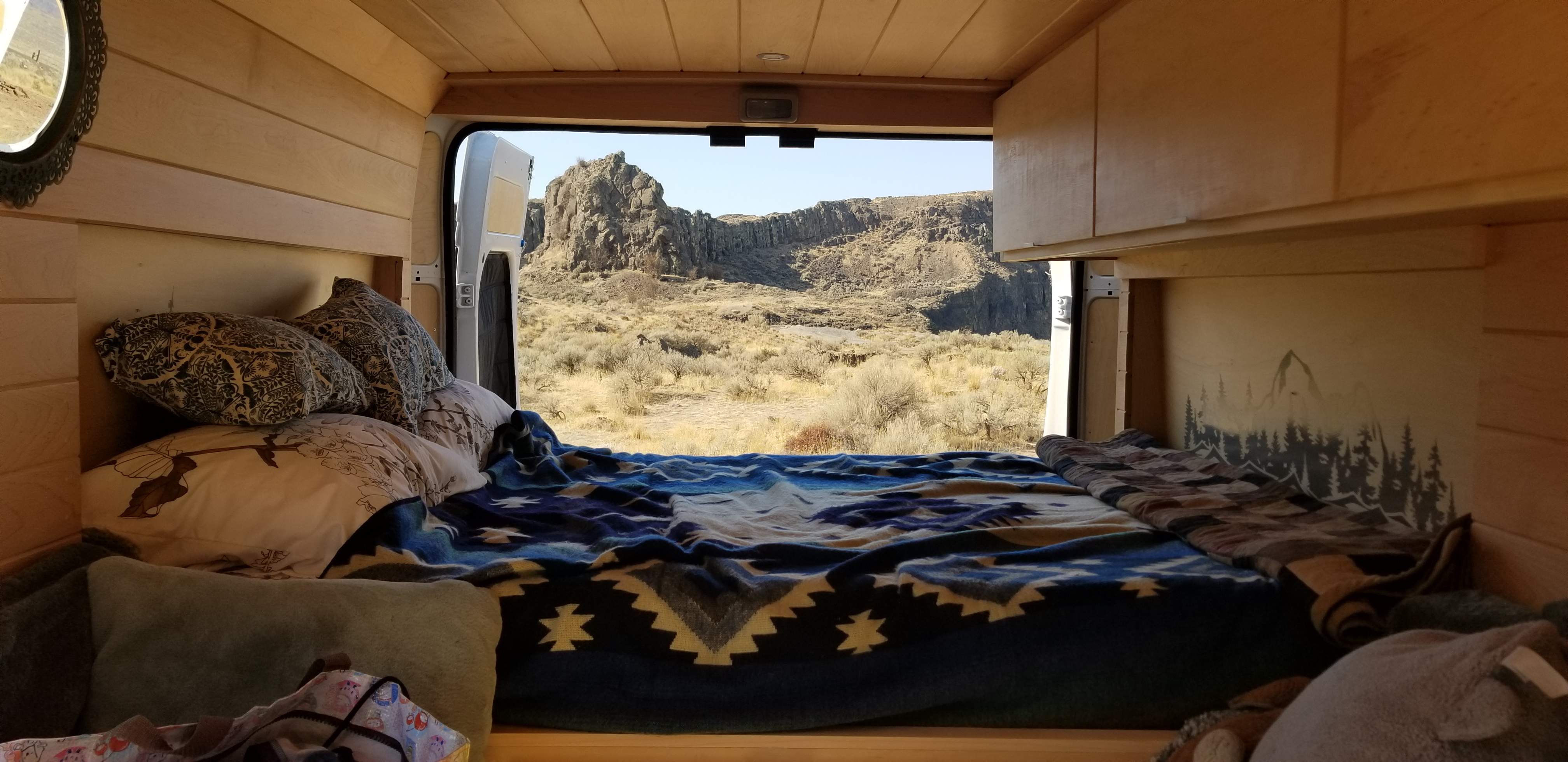 Cozy RV rental bedroom overlooks desert cliffs. Wood-paneled interior, comfortable bed with patterned blankets, and mountain mural.. Dodge Promaster, high roof 2020