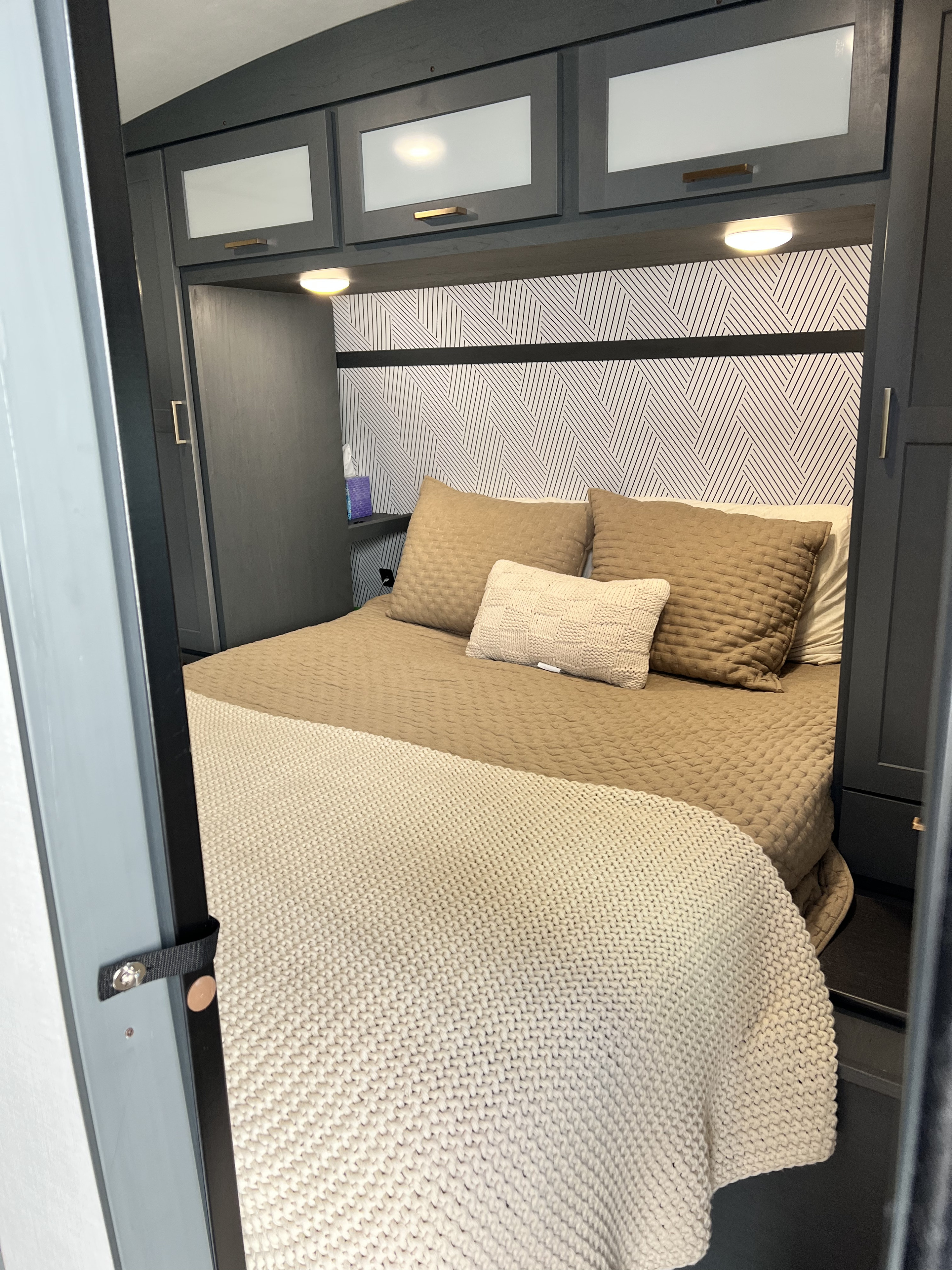 Modern RV rental bedroom featuring a cozy bed with neutral bedding, overhead storage cabinets, and stylish geometric wallpaper.. Keystone RV Bullet Ultra Lite 2022