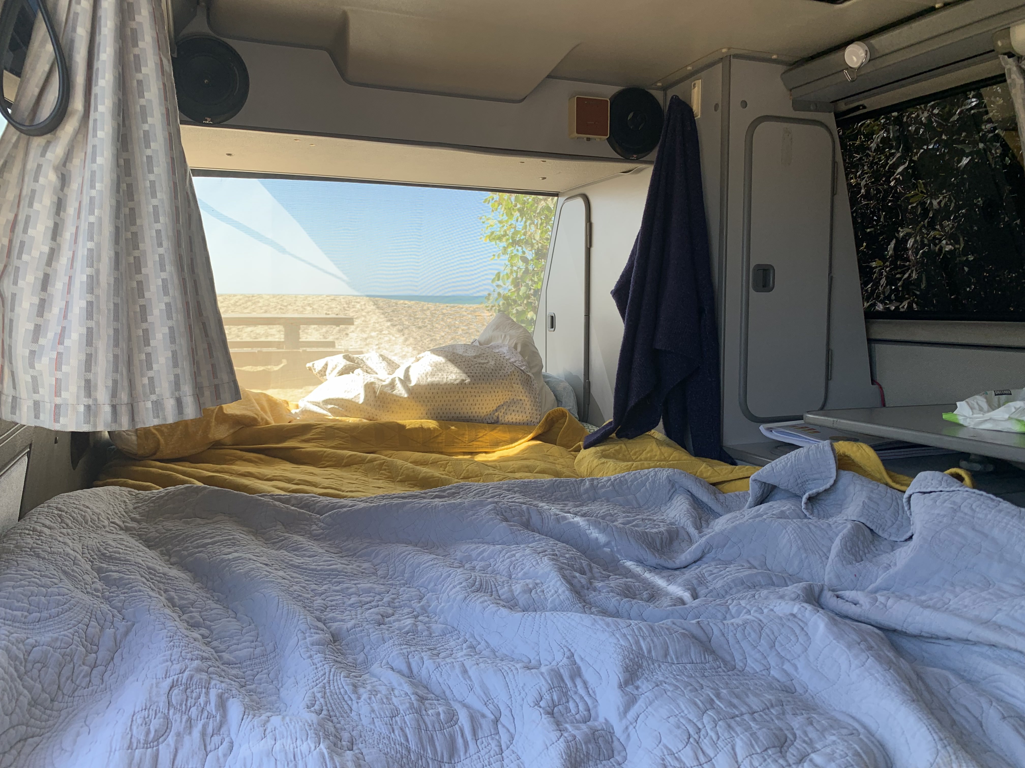 RV rental's comfy bed with blue & yellow blankets, facing a sunny beach view. Speakers & storage add to the cozy interior.. Volkswagen Westfalia 1988