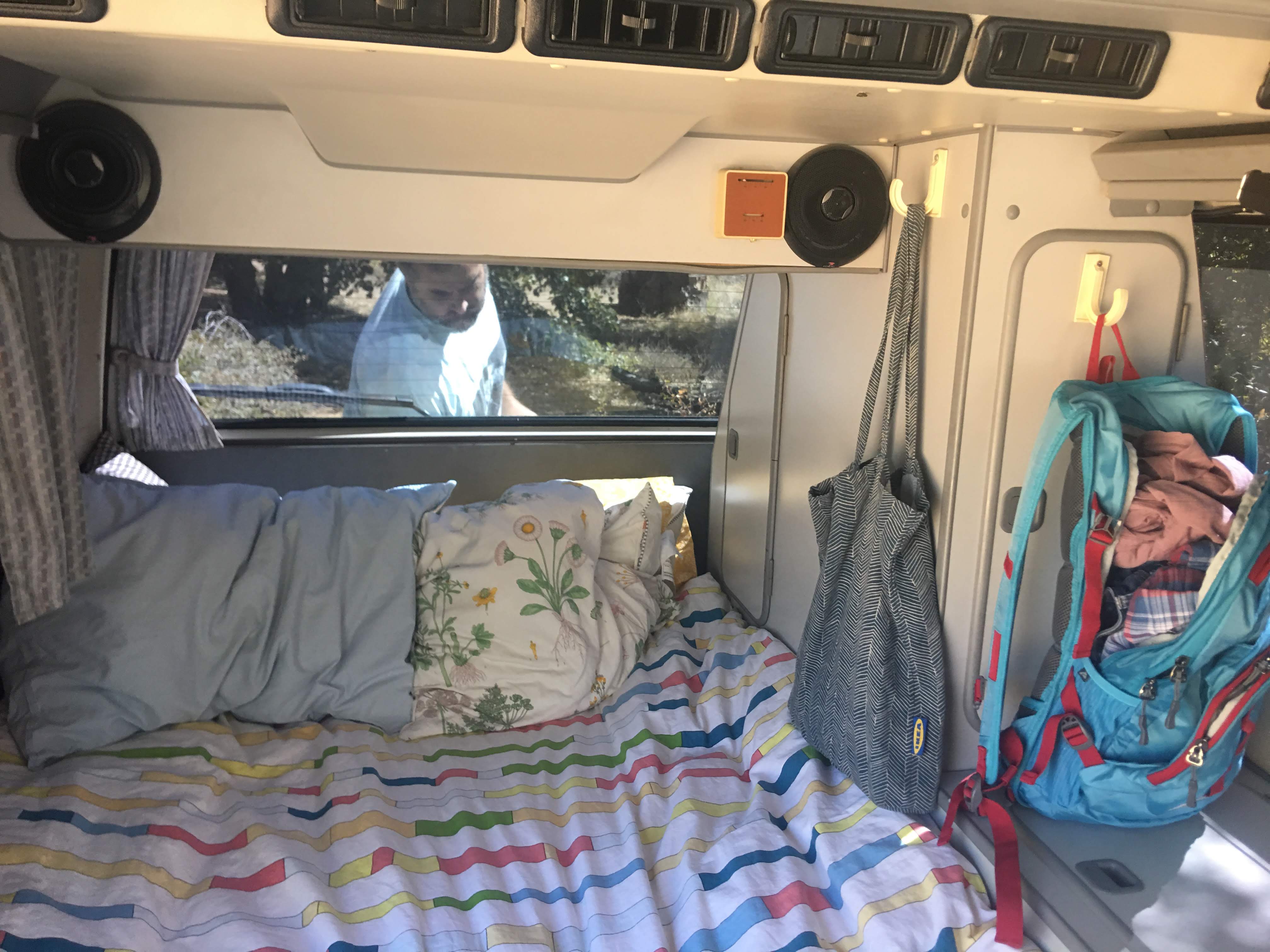 Cozy RV rental interior featuring a colorful bed, outdoor view with a person, and travel bags hanging on hooks.. Volkswagen Westfalia 1988