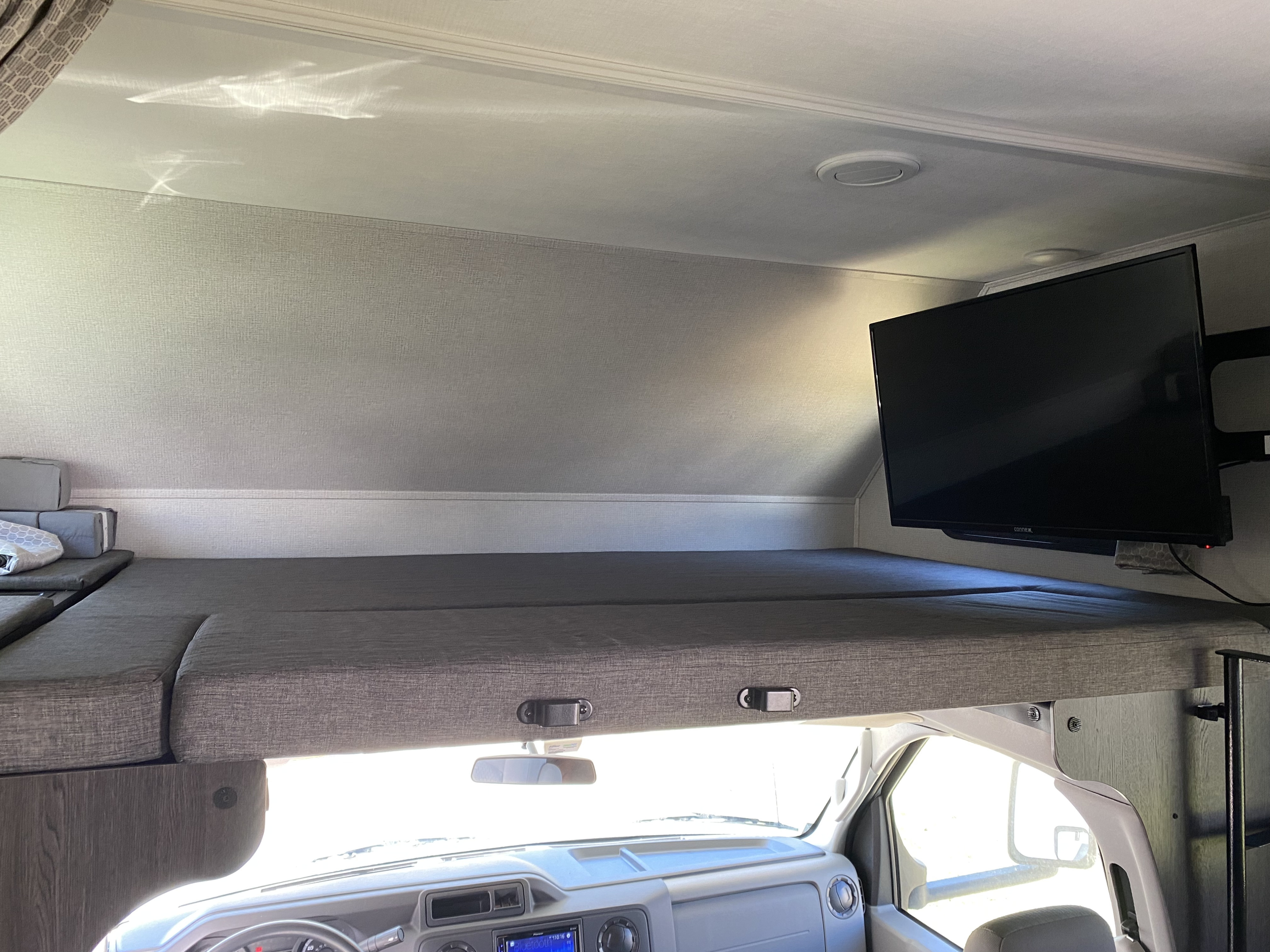 Inside an RV rental: a spacious over-cab bunk bed, mounted TV, and the bright dashboard of the motorhome cab.. Forest River Sunseeker 2021