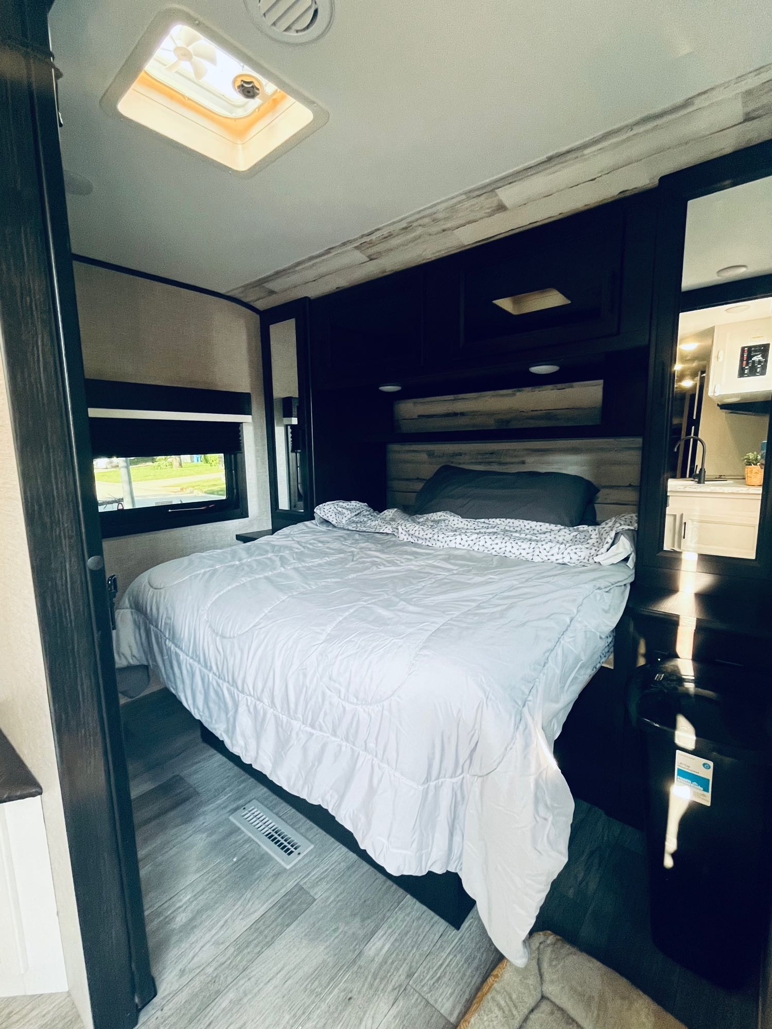 RV rental bedroom with a comfortable bed, overhead vent, window, and dark wood decor. A mirror reflects the kitchen area.. Jayco Jay Feather 2022