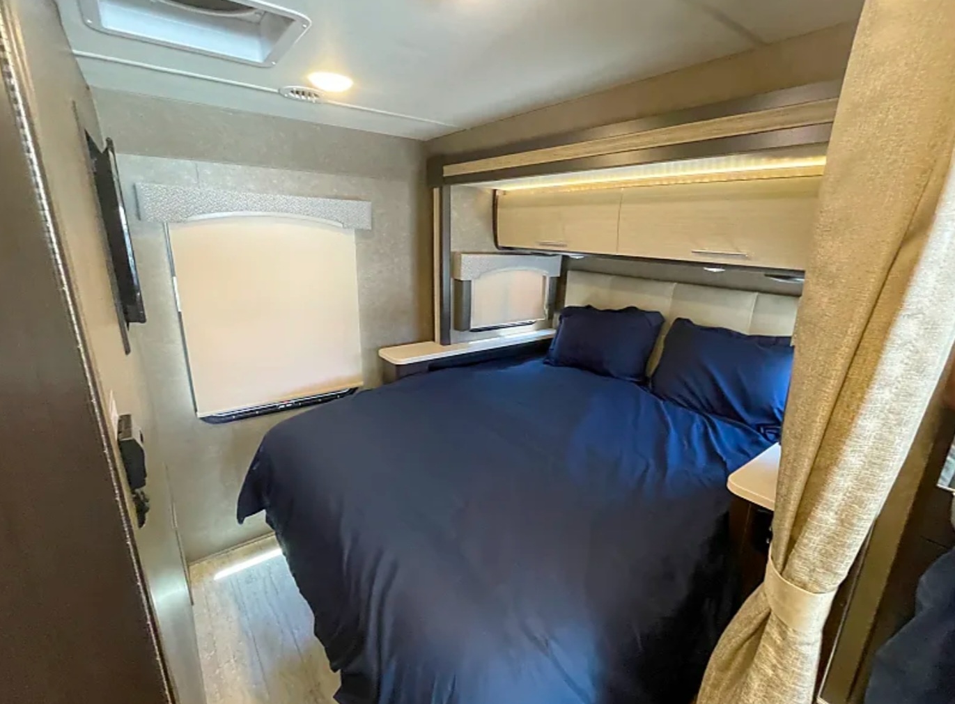 RV rental bedroom with a comfy dark blue bed, lighted overhead cabinets, and a side window for a cozy stay.. Thor Vegas 2018