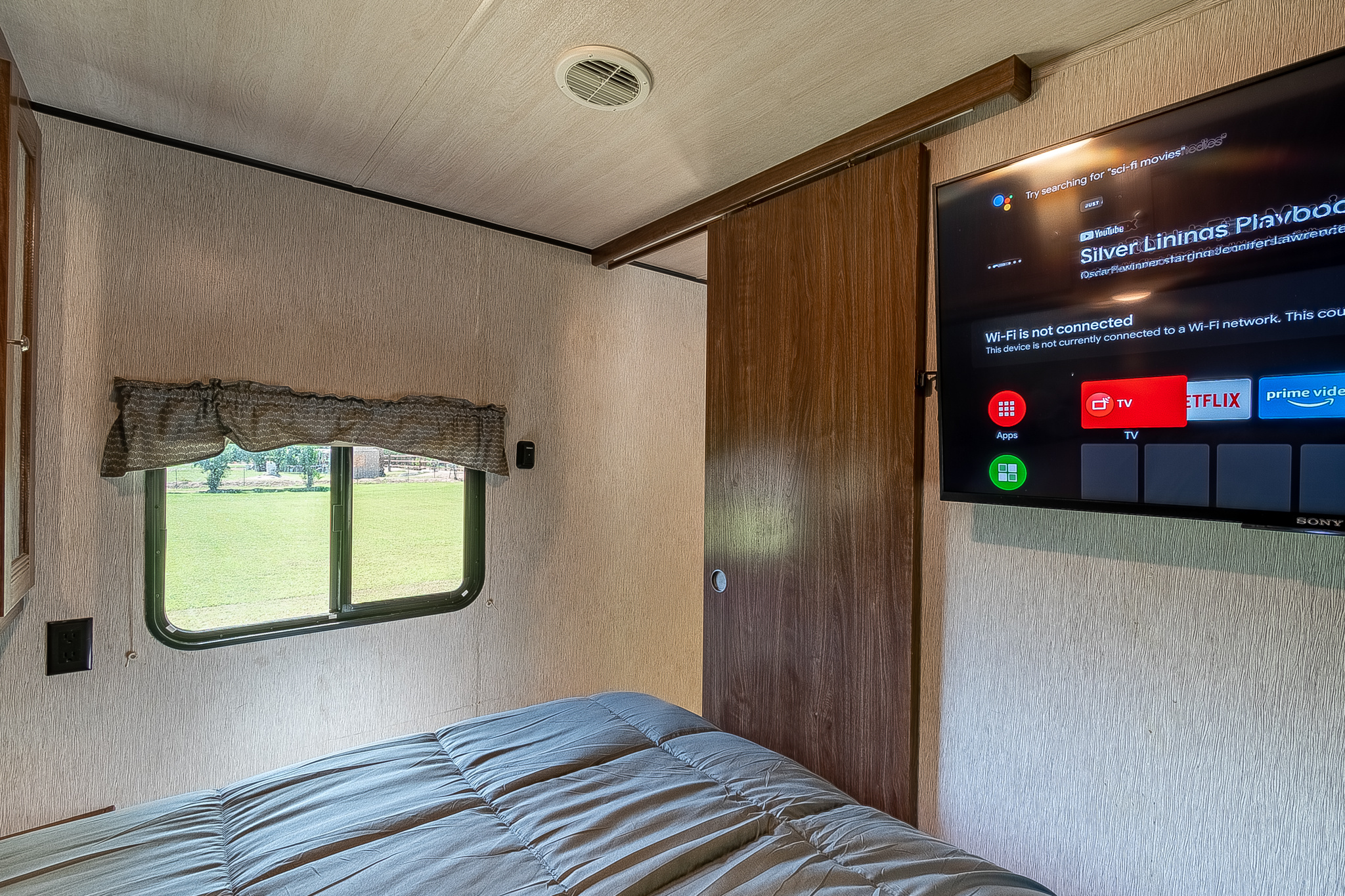 Inside an **RV rental**, a cozy bedroom features a gray bed, a window with a lush outdoor view, and a TV with app options.. Heartland RVs Pioneer 2018