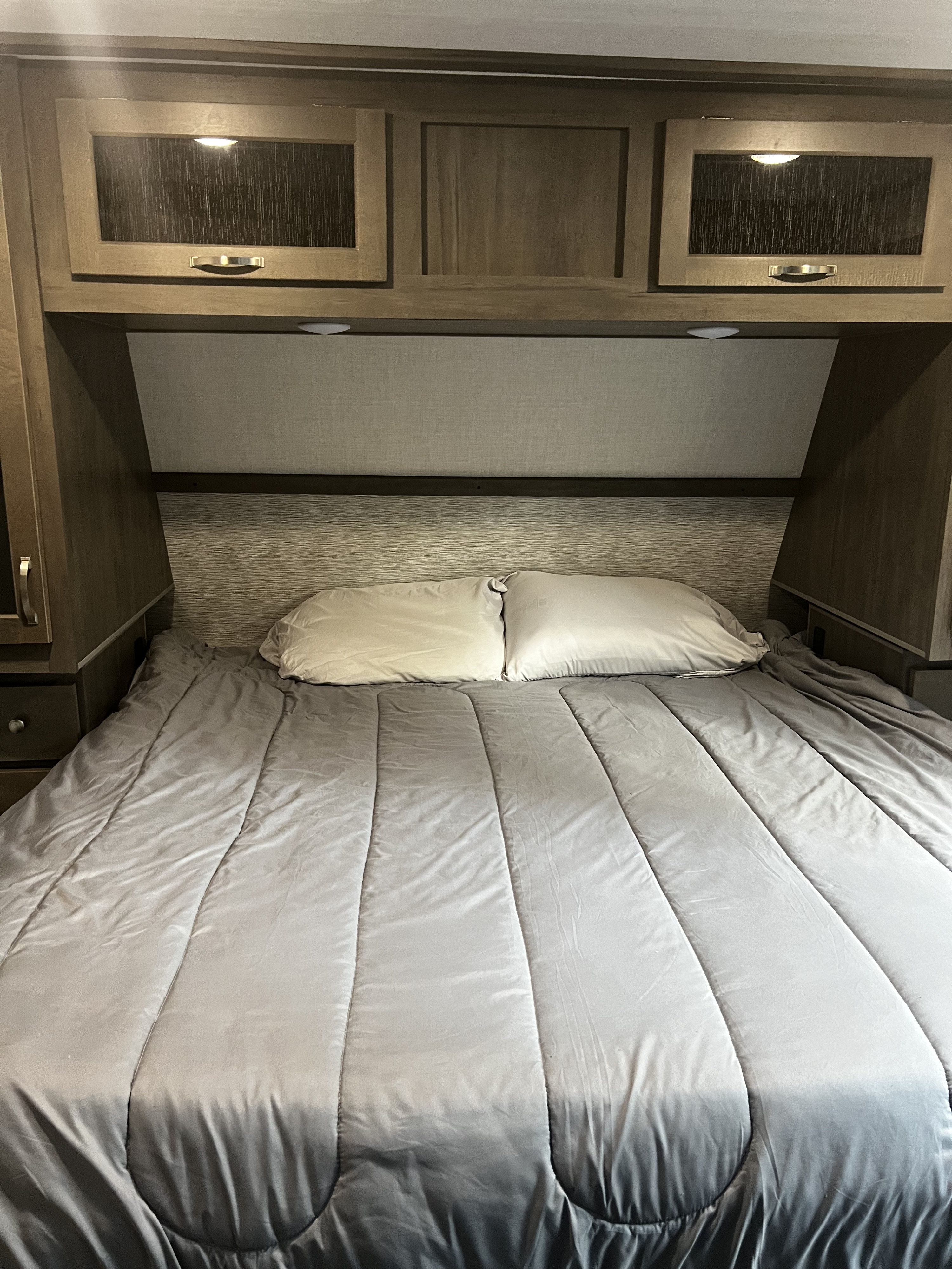A cozy RV rental bedroom. The bed has a grey comforter & white pillows. Wooden cabinets & lighting are above the headboard.. East to West Alta 2021