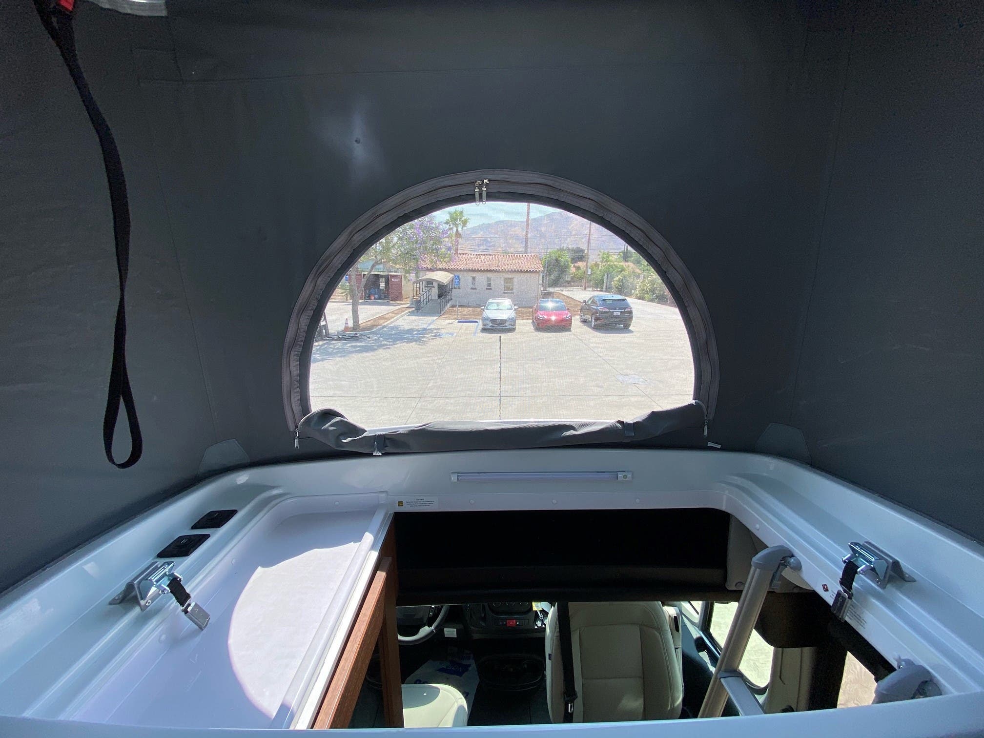 Experience an RV rental! Inside a camper van's pop-top, a bed area with an oval window overlooks a lot, building & mountains.. Roadtrek Roadtrek Class B 2021