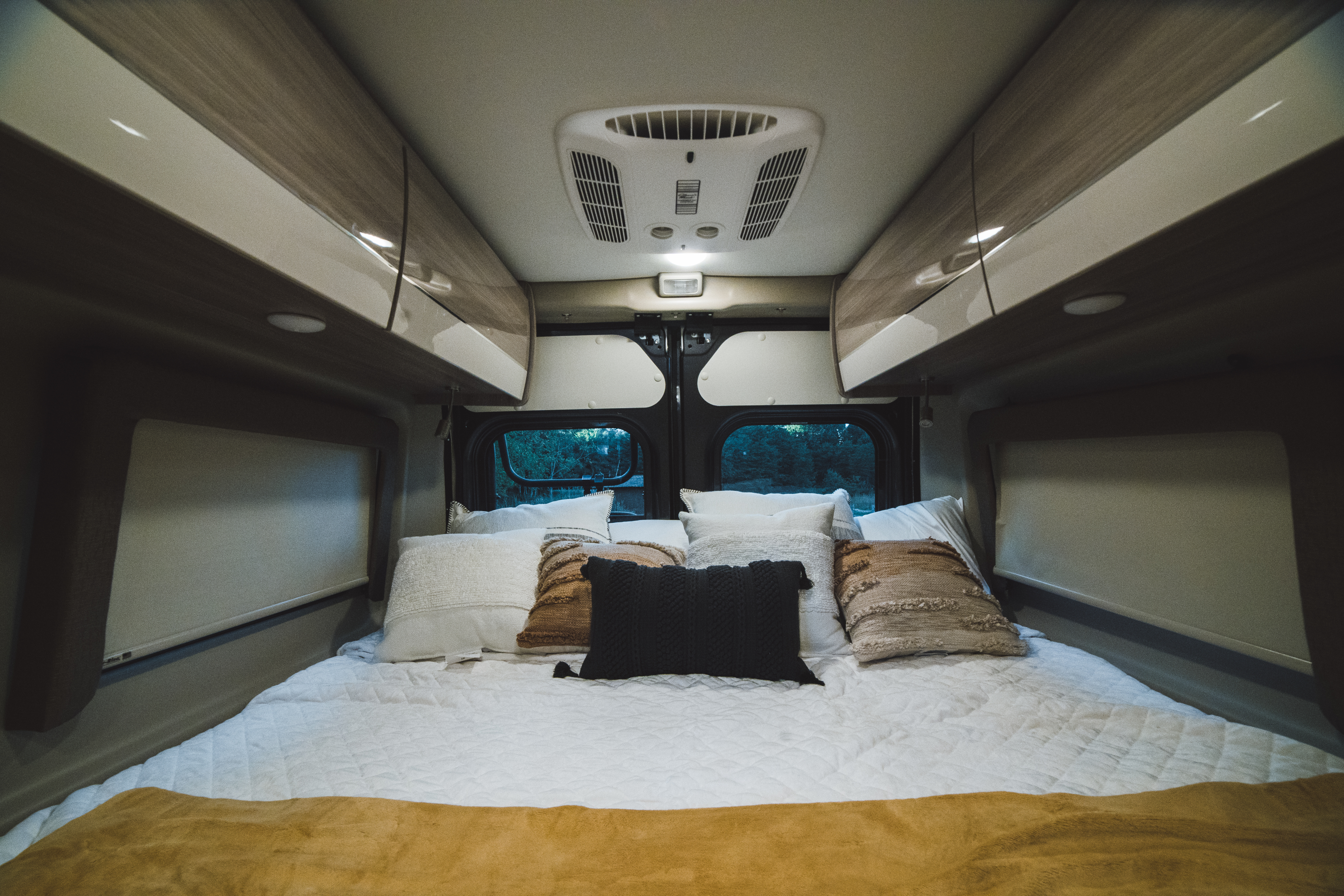 Cozy RV rental bedroom featuring comfortable bedding, overhead cabinets, a ceiling AC unit, and rear window view.. Thor Sequence 2022
