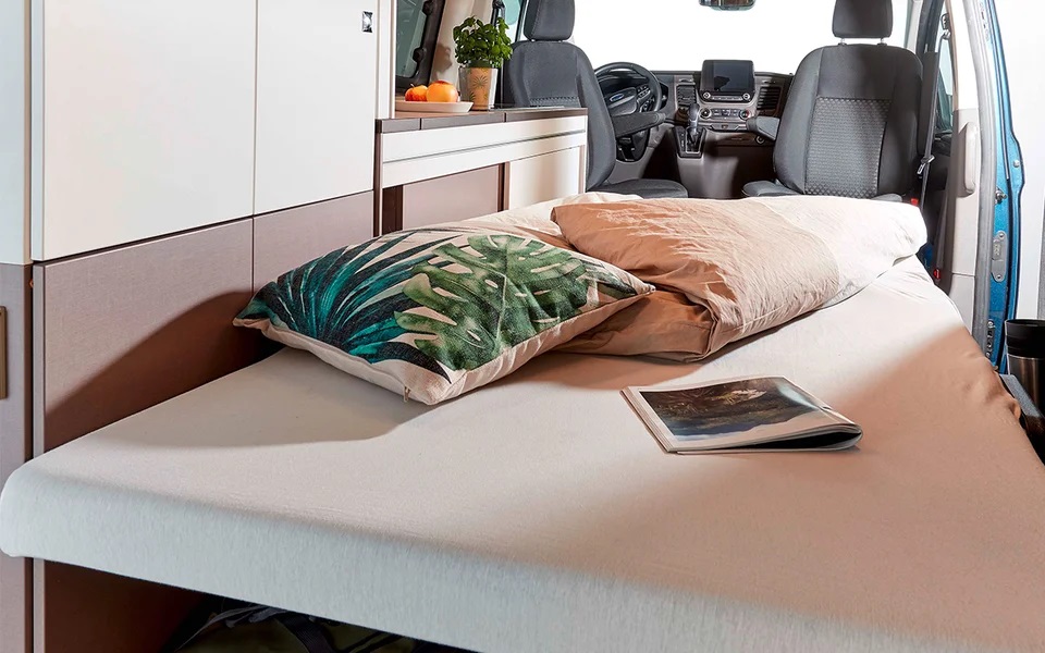 Cozy RV rental interior featuring a comfortable bed with a tropical pillow and magazine. Perfect compact living for travel.. Bürstner COPA 500 2023