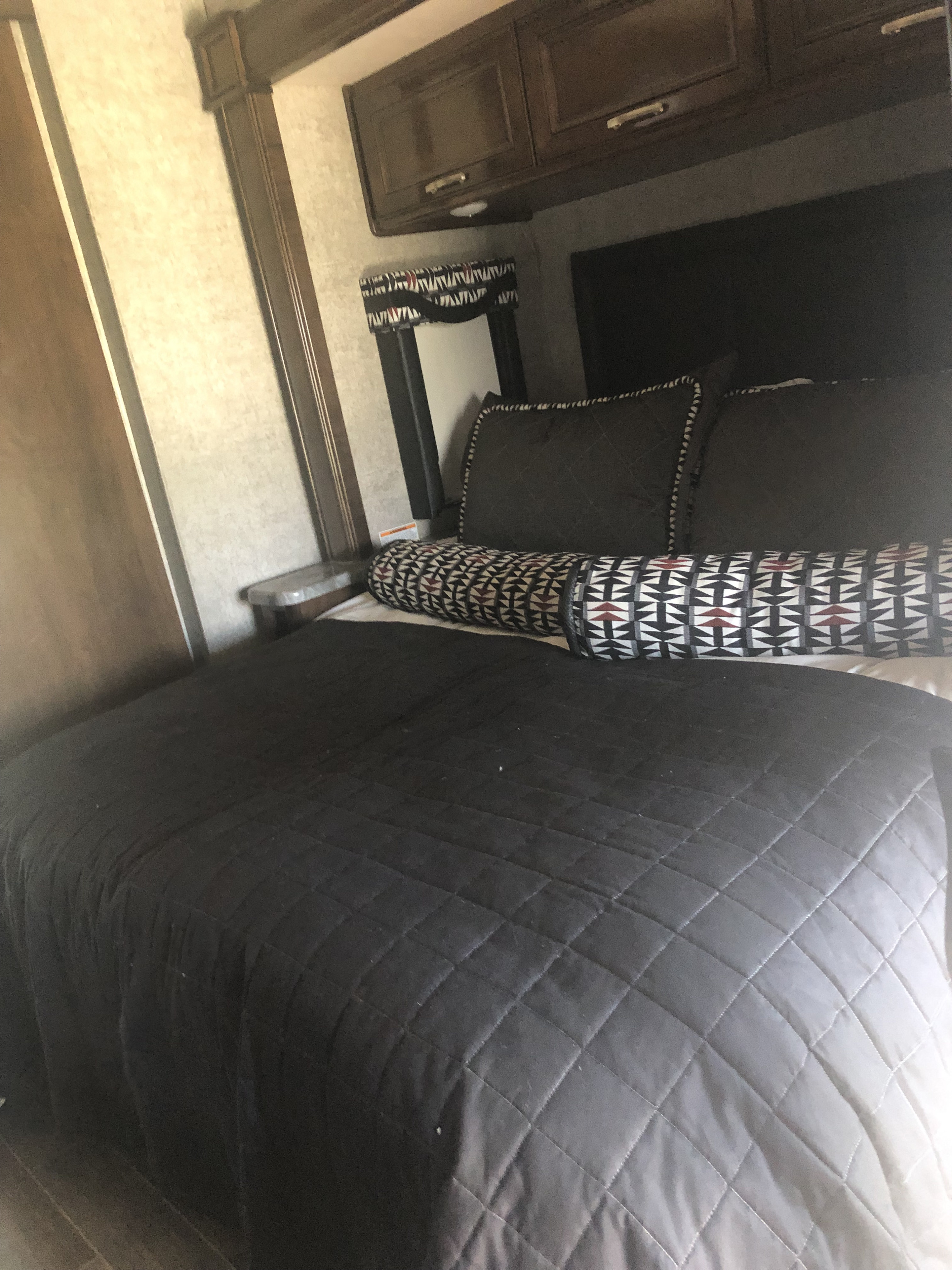 RV rental bedroom featuring a bed with dark quilted bedding, patterned pillows, and overhead storage.. Thor Outlaw 2020